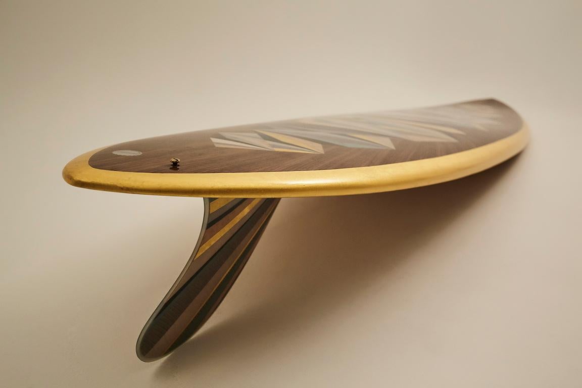 Il s'agit d'une planche de surf unique et personnalisée, dotée d'un pont en marqueterie incorporant des feuilles d'or 24 carats, conçue et fabriquée à la main par le studio w o o d p o p, spécialisé dans la marqueterie et les incrustations. 

Le