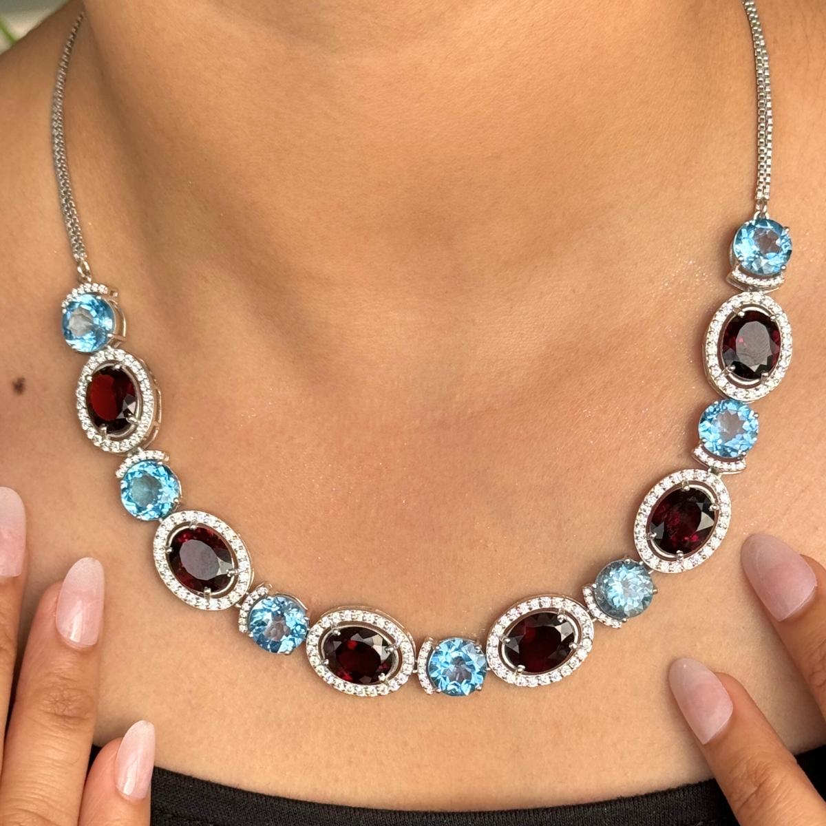 Diese zweifarbige Granat- und Blautopas-Edelsteinkette mit CZ-Halo ist eine atemberaubende Halskette aus 925er Sterlingsilber, die eine luxuriöse Kombination aus tiefroten Granat- und lebhaften Blautopas-Edelsteinen präsentiert. Jeder ovale Granat