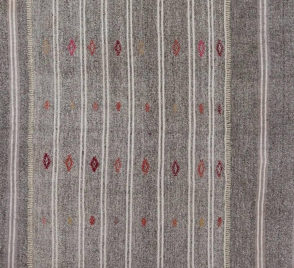 A large, simple, minimalist Turkish flat-weave/kilim patchwork rug made up of hand-woven and hand-stitched vintage kilim pieces that feature stripes and colorful burdock motifs against a grainy looking texture of the background, in gray and white.