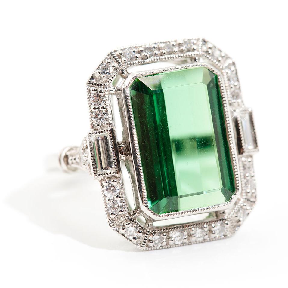 Forged in platinum is this art deco inspired halo ring featuring a breathtaking 9.96 carat bright green modified emerald cut natural tourmaline, embellished with total of 0.70 carats of sparkling round brilliant cut and baguette cut diamonds. We