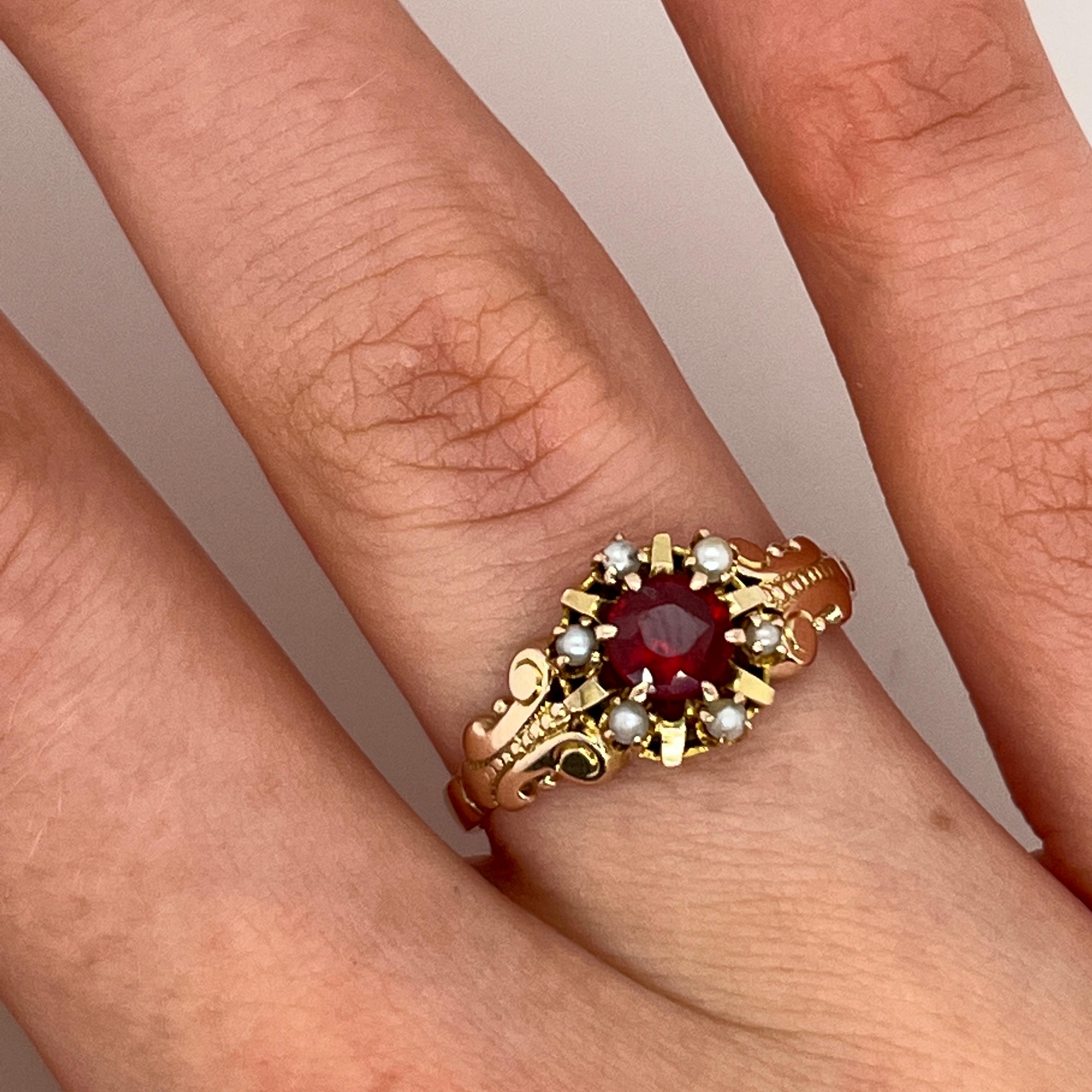 Realizzato in caldo oro giallo 9ct, questo affascinante anello in stile vintage presenta un vibrante rubino a taglio rotondo al centro, accentuato da delicate perle di semi disposte a raggiera. La combinazione di un rosso ricco e di una morbida