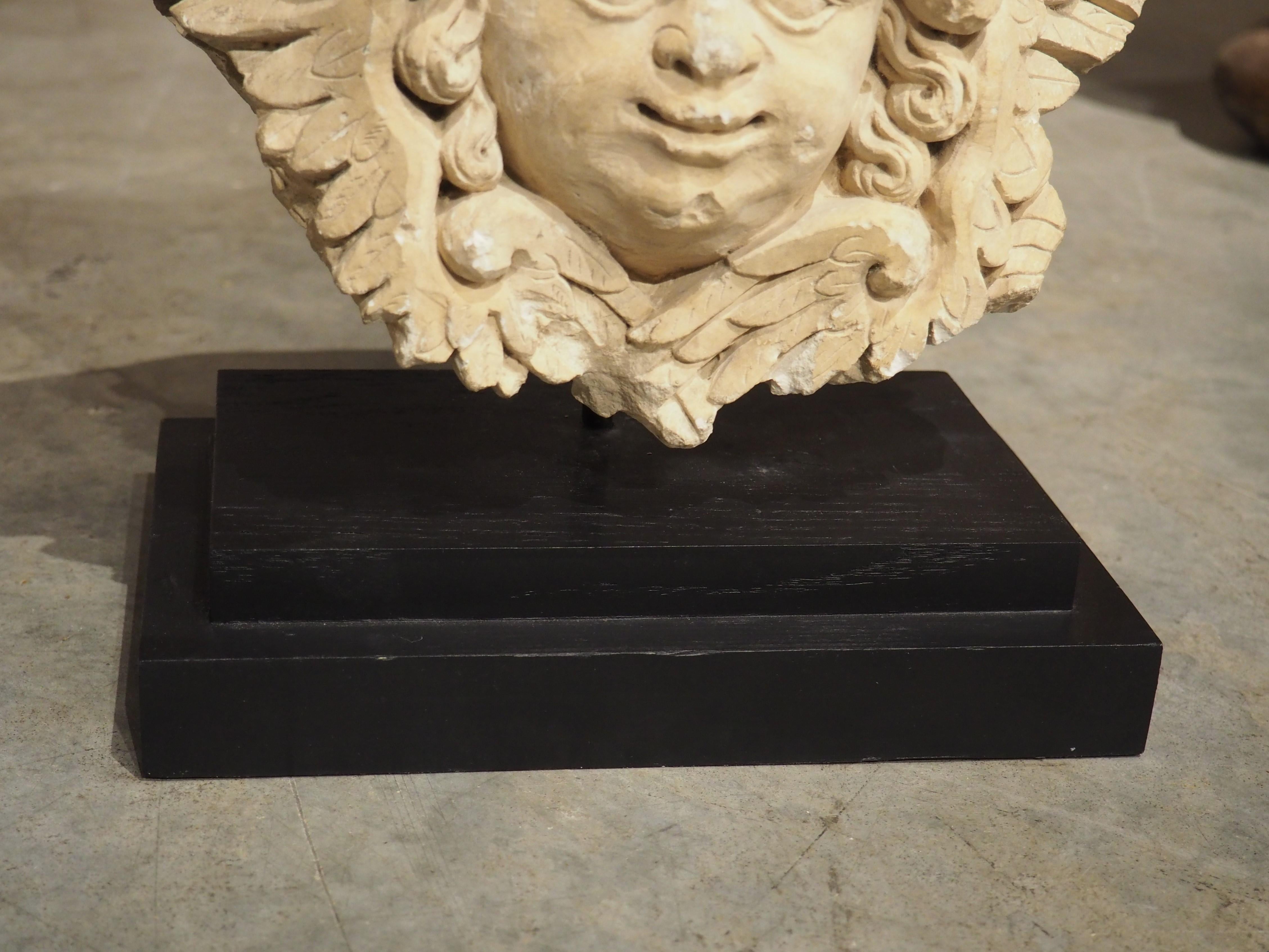 Originally part of a larger architectural from a French building in the 1600s, this hand-carved limestone fragment of a winged angel has been mounted more recently to a painted black stand with a metal arm. The plinth has a stepped molding and a