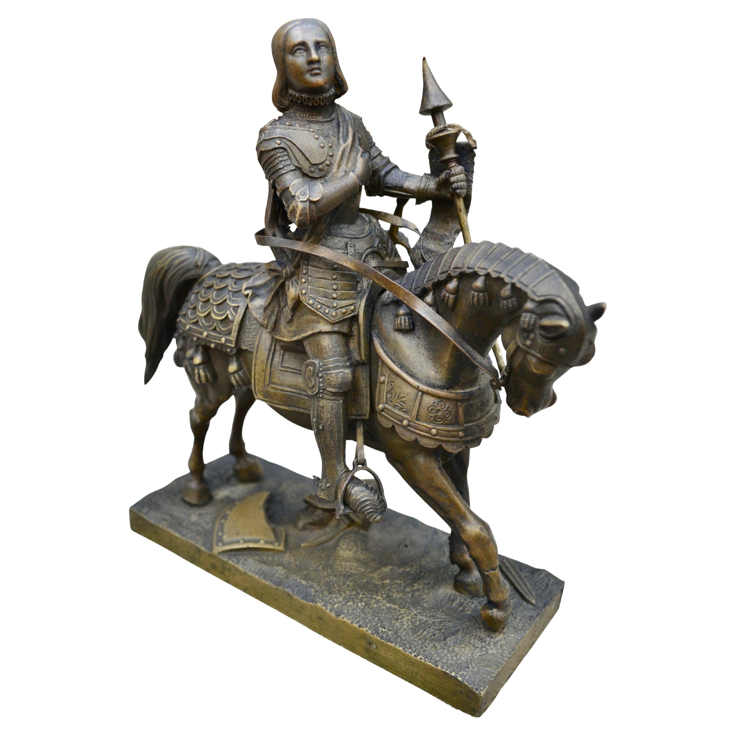 An small but exquisitely cast and highly detailed bronze statue of St Joan of Arc in full body  battle armour including the horse with one hand holding a standard and the other hand resting on her heart. Her sword is still in its sheath hanging by
