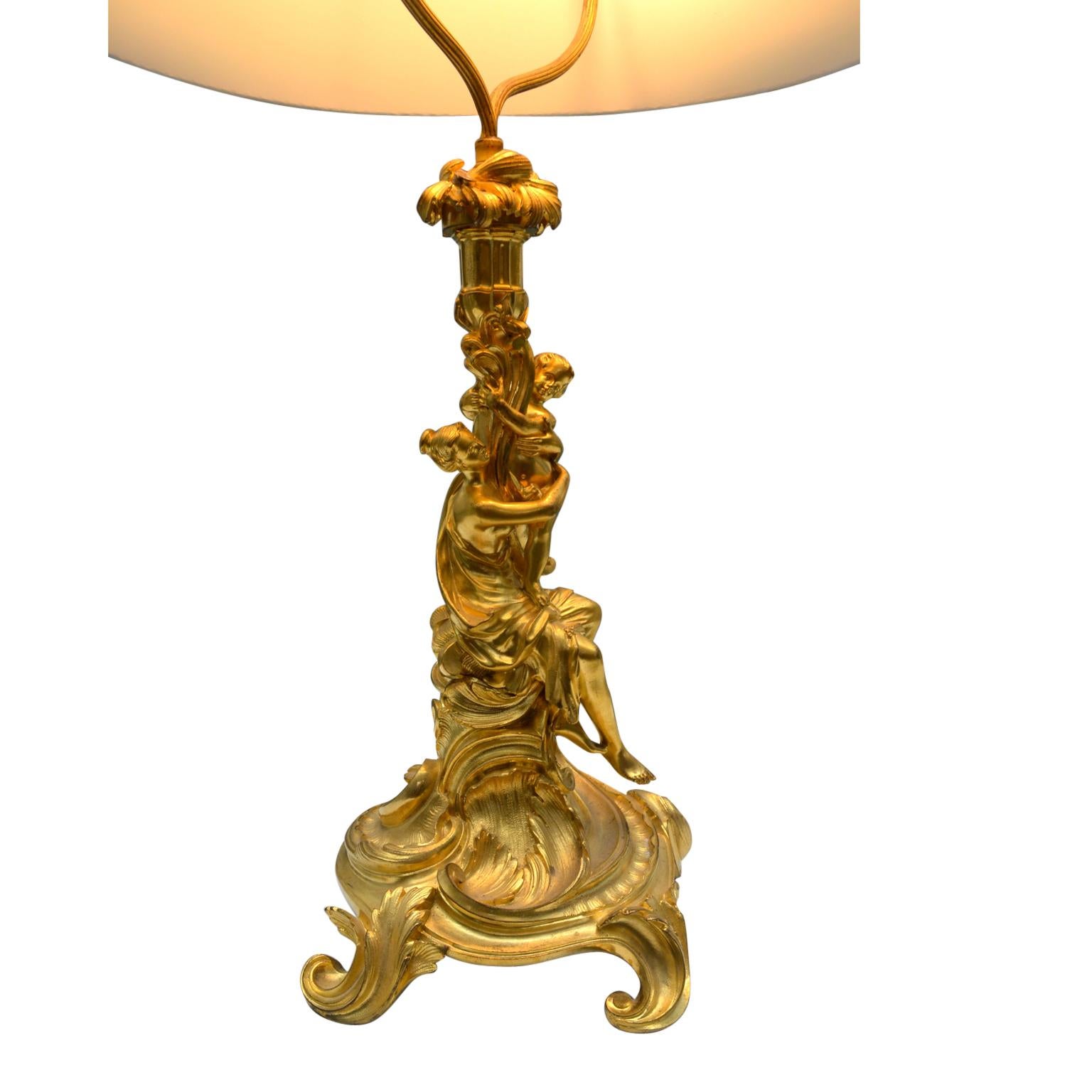 A French gilded bronze figural lamp featuring a standing classically draped lady with a cupid in her arms who further holds aloft a cornucopia containing the socket; the whole resting on a circular Rococo styled base.