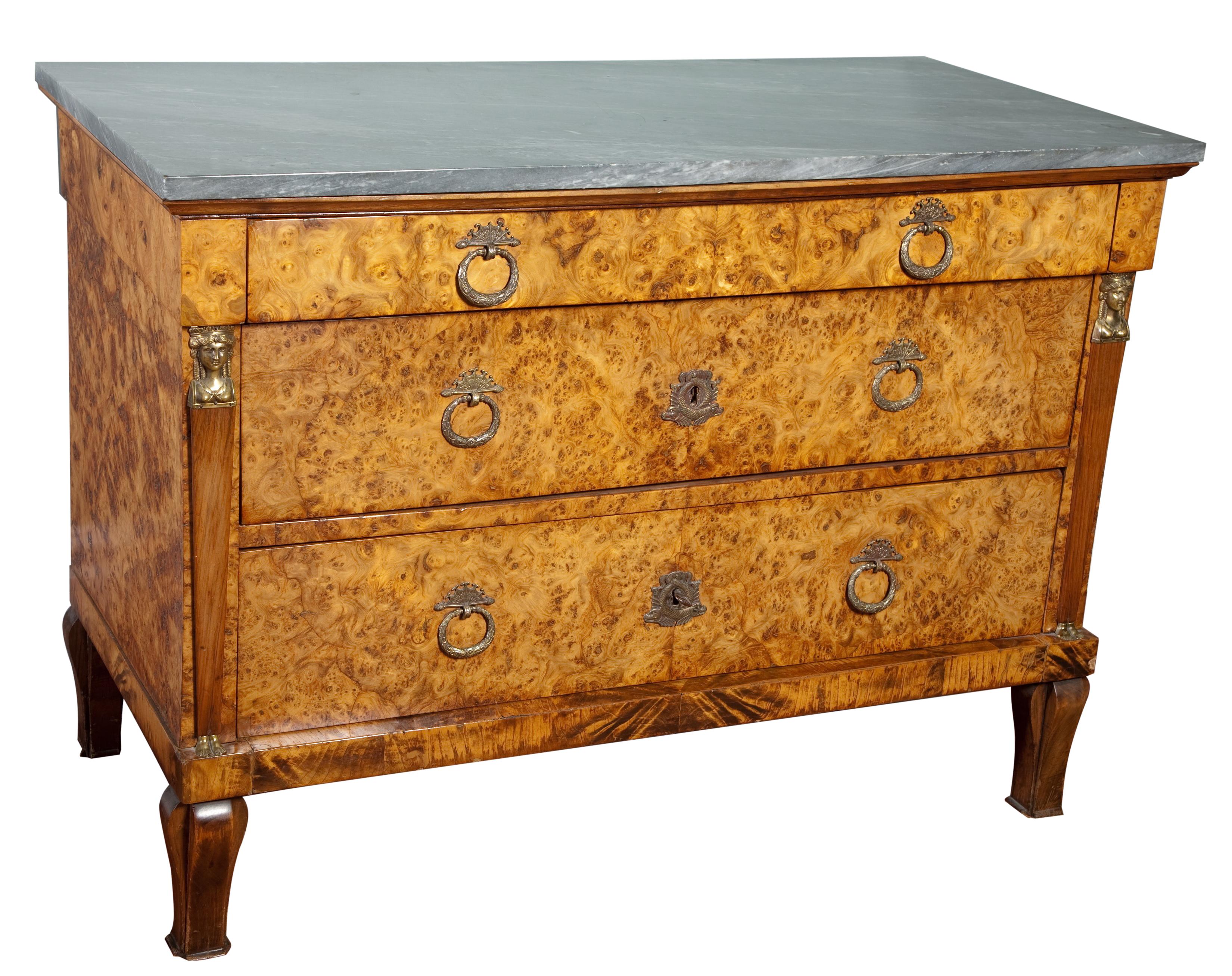 A Swedish Empire three-drawer commode chest veneered in burled bird's-eye elm emblematic of  Baltic  furniture in the 18th-19th centuries, the rectangular side columns mounted with brass Egyptian heads and feet, topped with a slab of bleu turquin