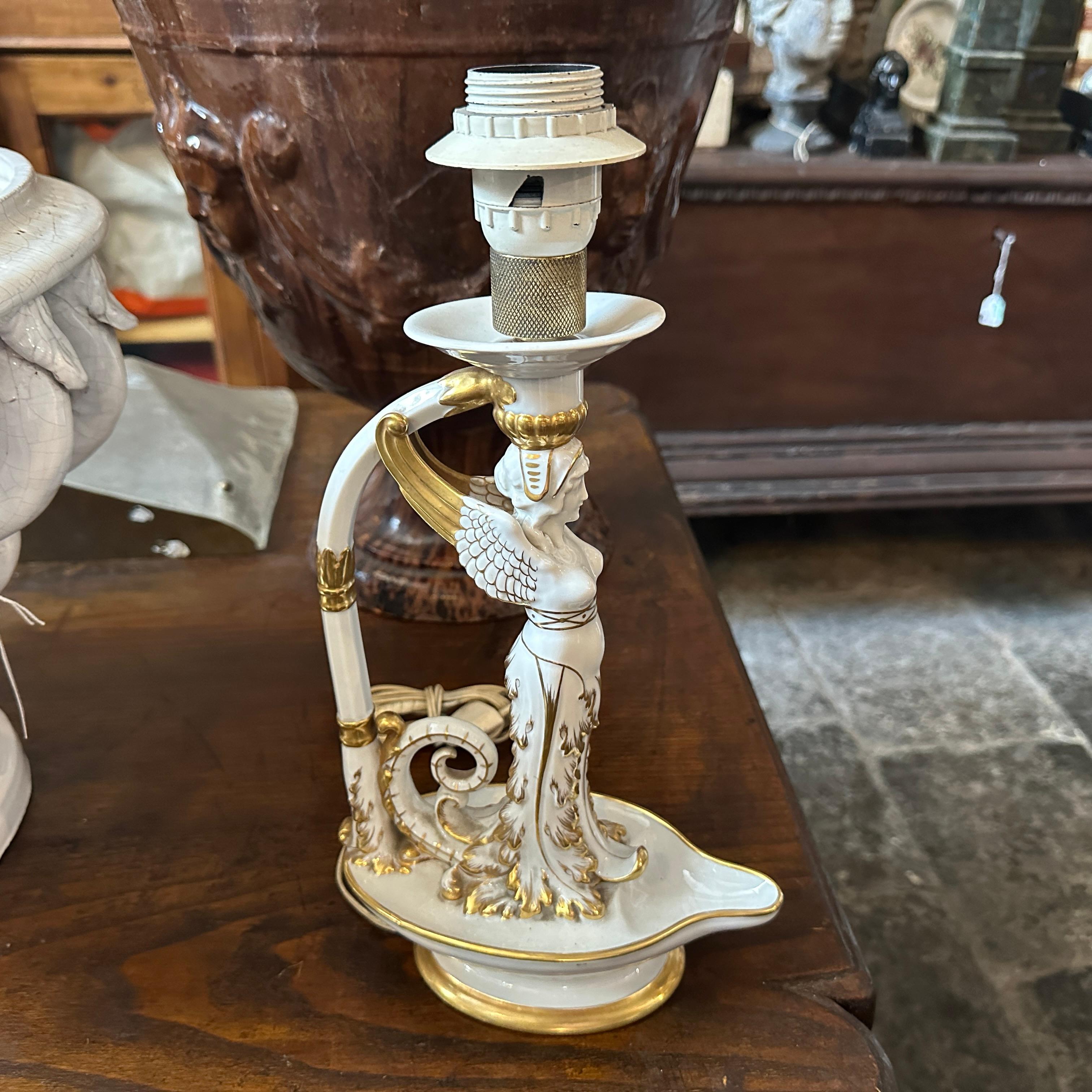The lamp features a white and gold color scheme, with intricate details including a winged female figure and elaborate scrollwork. The lamp has been manufactured in the early 1900s, exemplifying the ornate and classical designs typical of that era.