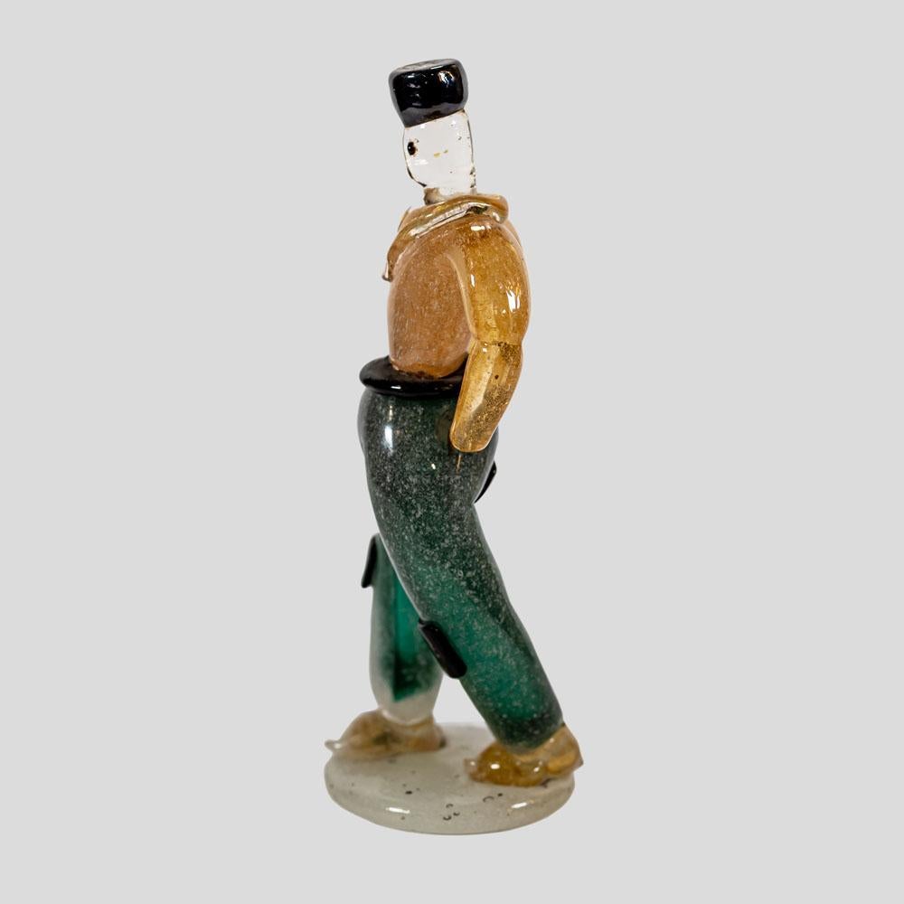 A rare 1930s Murano blown glass figure of a young man with a black hat, gold dust top and green trousers, clear and gold submerso shoe positioned on a off white round glass base.. This technique is blown glass fusion of gold, green black and clear