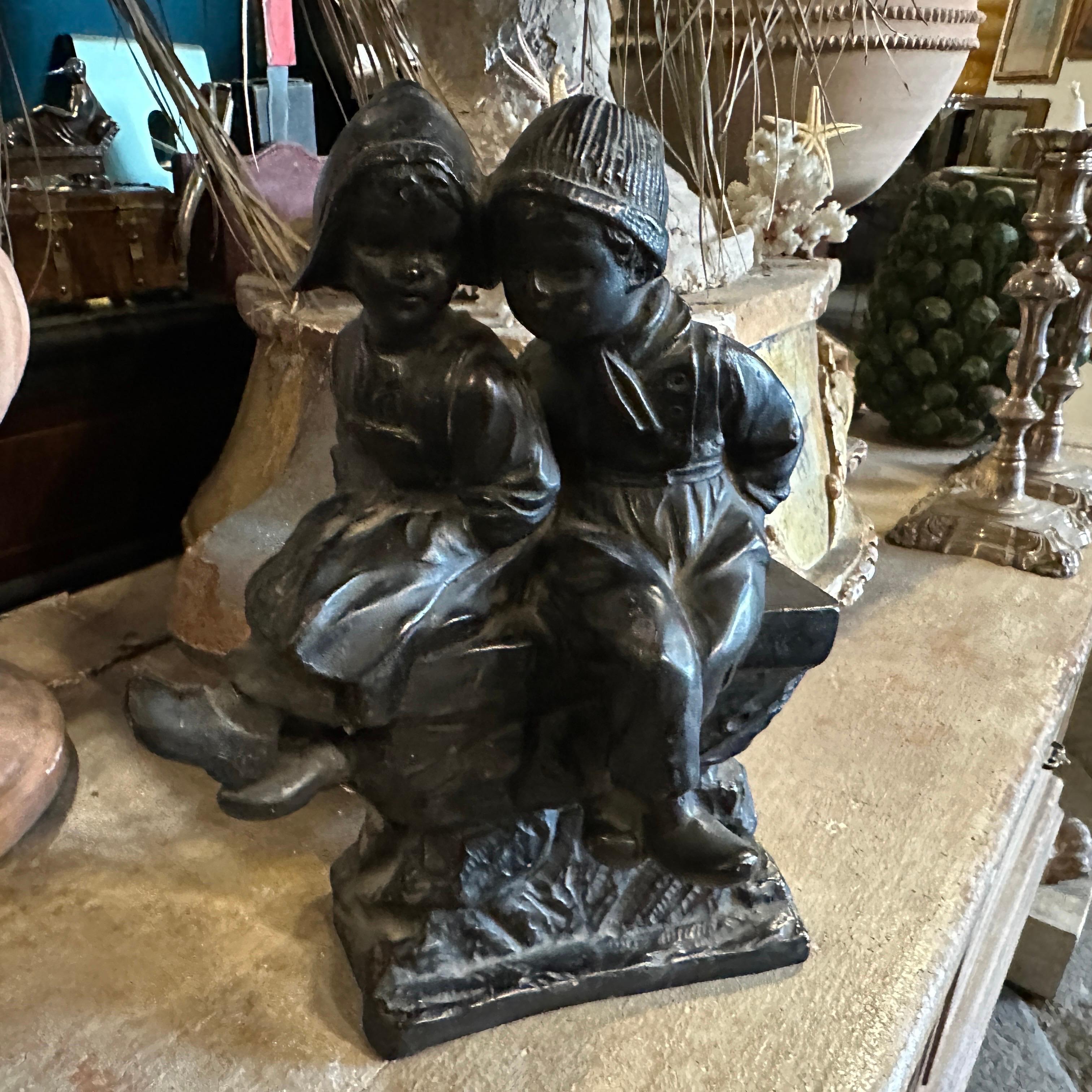 Il s'agit d'une figurine en terre cuite peinte en noir, datant des années 1950, représentant deux enfants néerlandais assis ensemble sur un banc. La sculpture présente un style d'art populaire européen traditionnel, avec des détails complexes dans