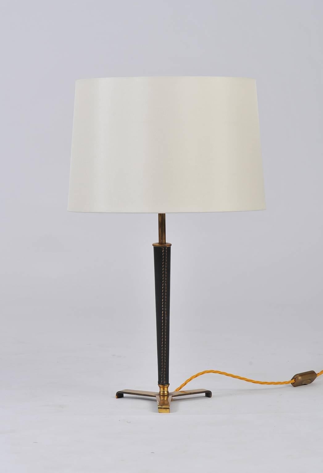 A brass and stitched black leather lamp, in the manner of Jacques Adnet, topped with an ivory silk tapered shade.
France, circa 1950

(Dimensions inclusive of the shade).