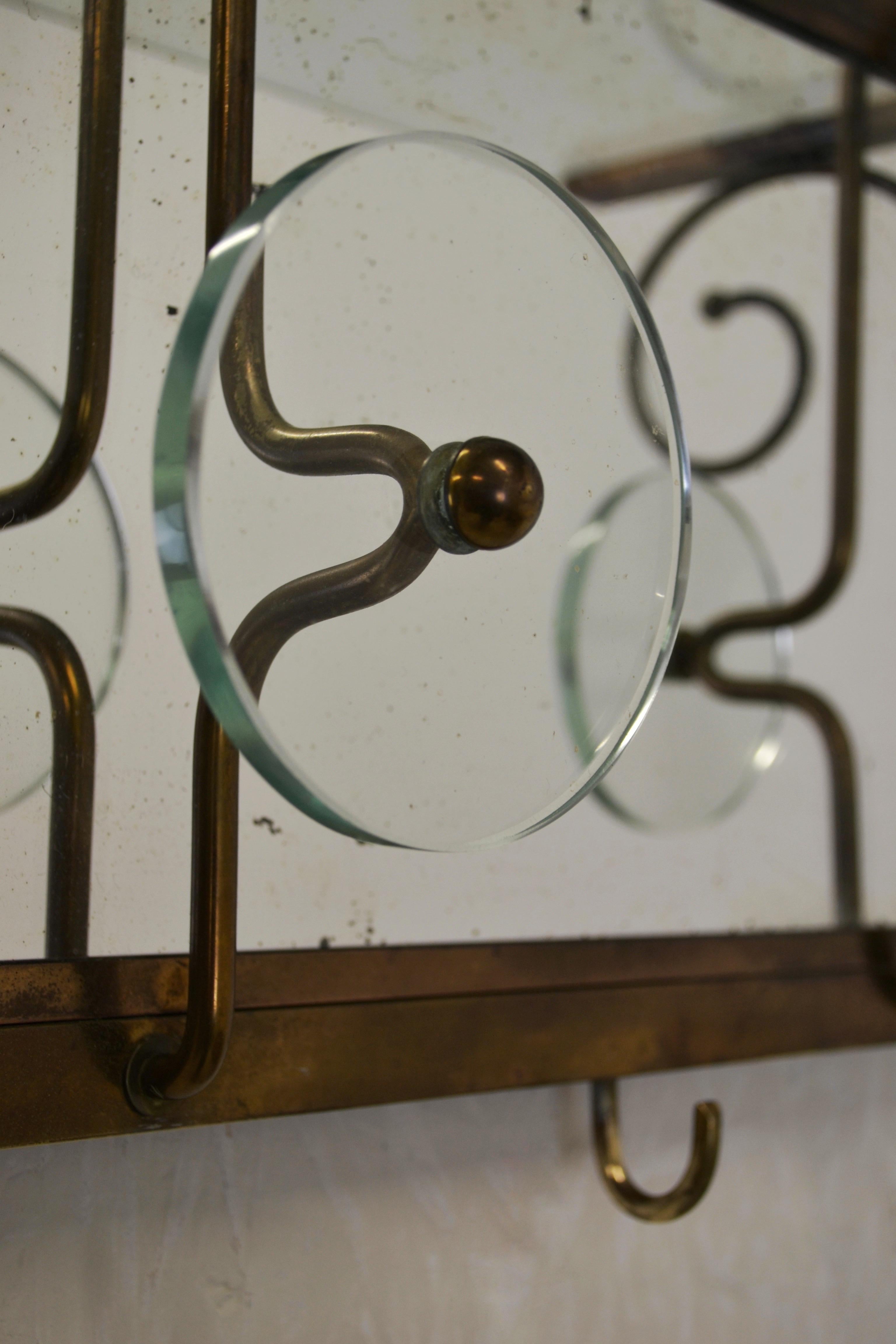 This 1950s Italian wall-mounted coat rack, inspired by the Fontana Arte style, stands out with its elegant design. The patinated brass frame surrounds an antiqued mirror, highlighting three oversized round glass knobs and two small brass hooks. The