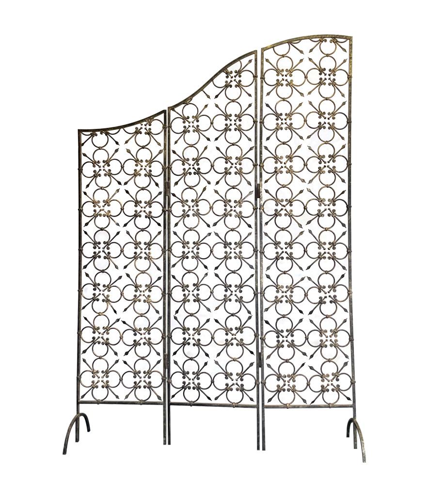 A 1950s Spanish wrought iron three panel screen with sloping curved top and intricate flower like detail, sitting on wrought iron feet, with gilt and black painted finish.