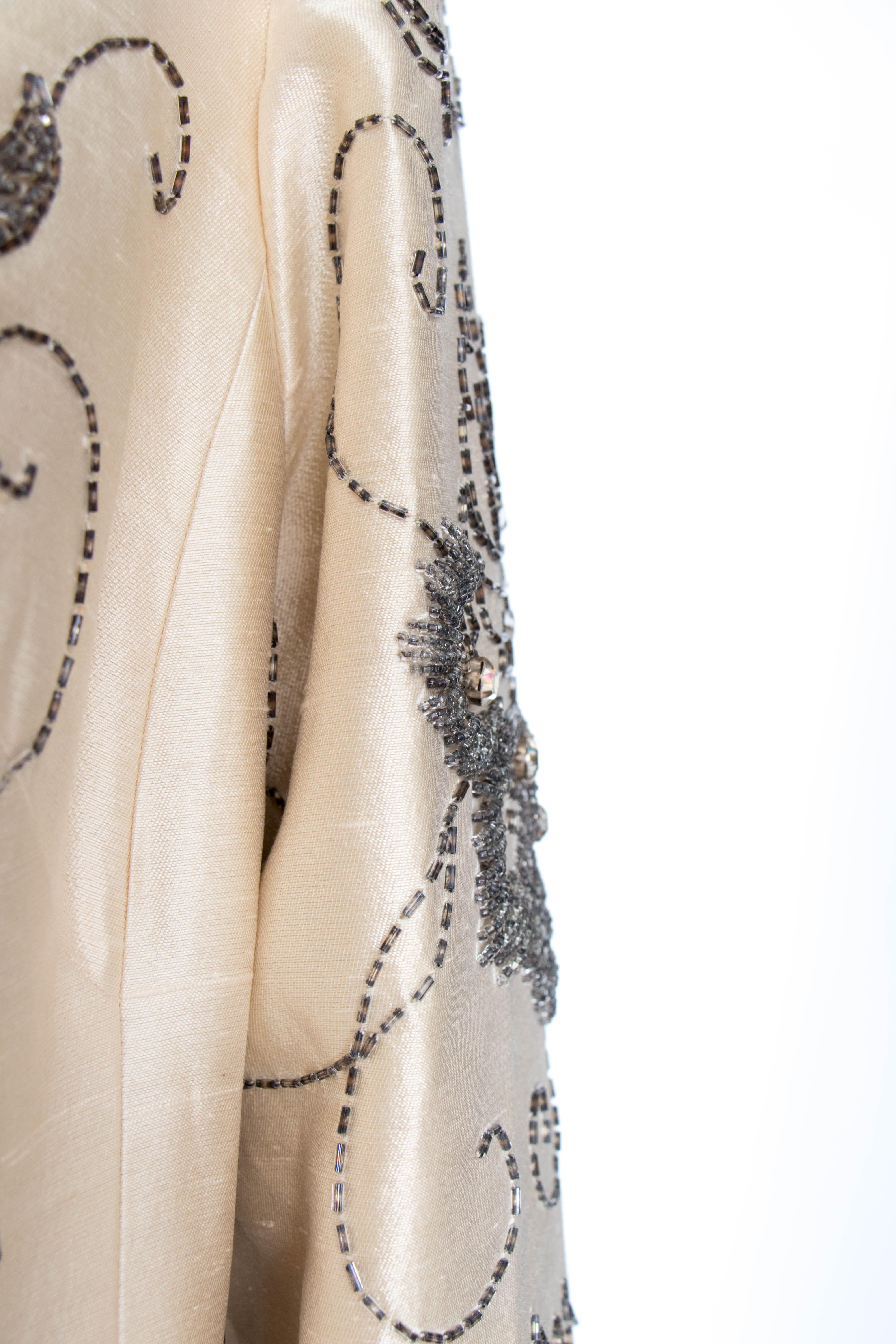 An incredible 1960s couture silk jacket with a round collar and a hidden front button closure. Intricate beading with silver beads and clear rhinestones are set in a floral pattern across the coat. The coat is fully lined.  

