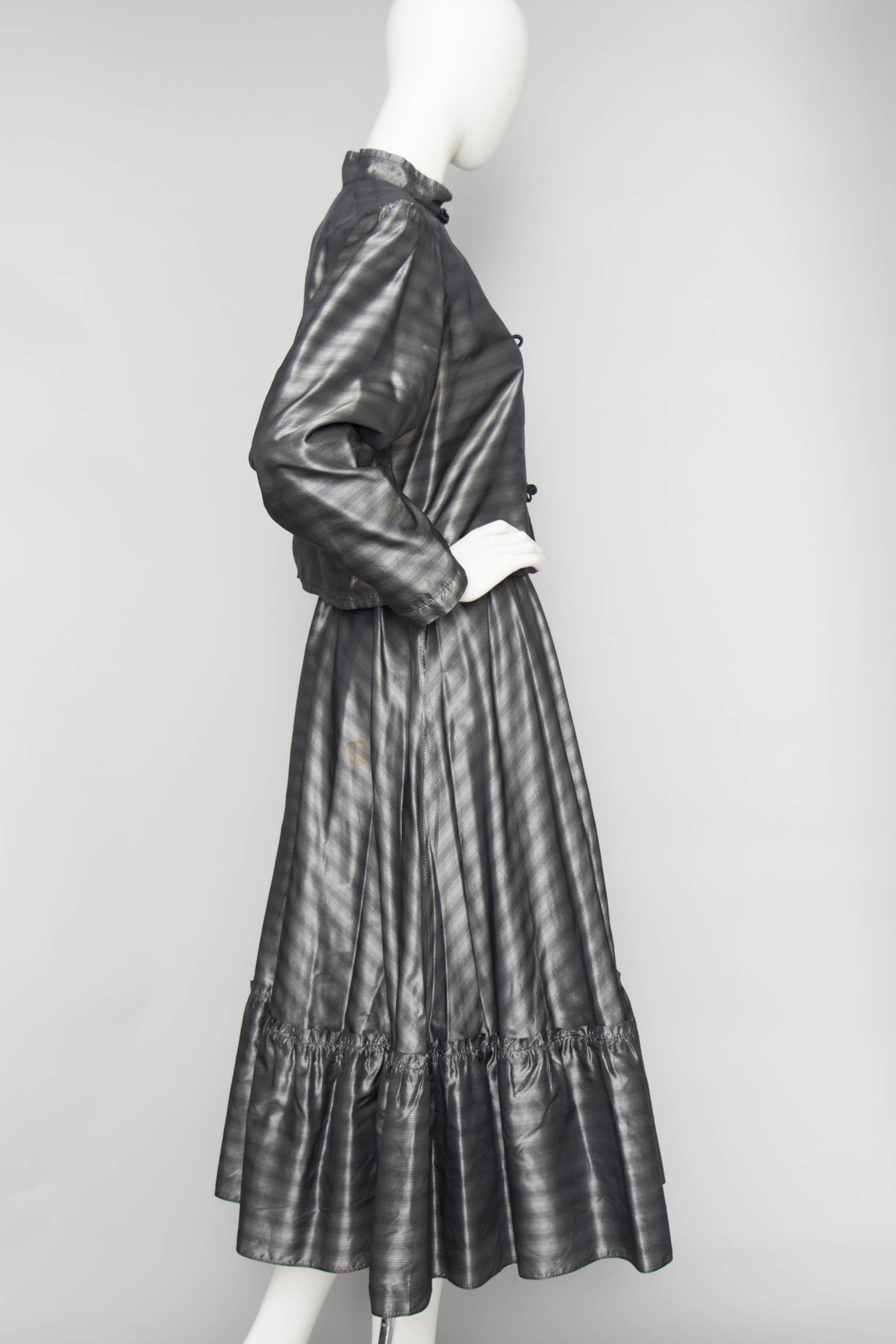 A 1970s Ted Lapidus silver taffeta ensemble consisting of a jacket and flared skirt with a ruffle hemline. and a blouse with puffed shoulders, tapered sleeves, a high collar, and knot buttons. The Item is fully lined.

The size of the ensemble