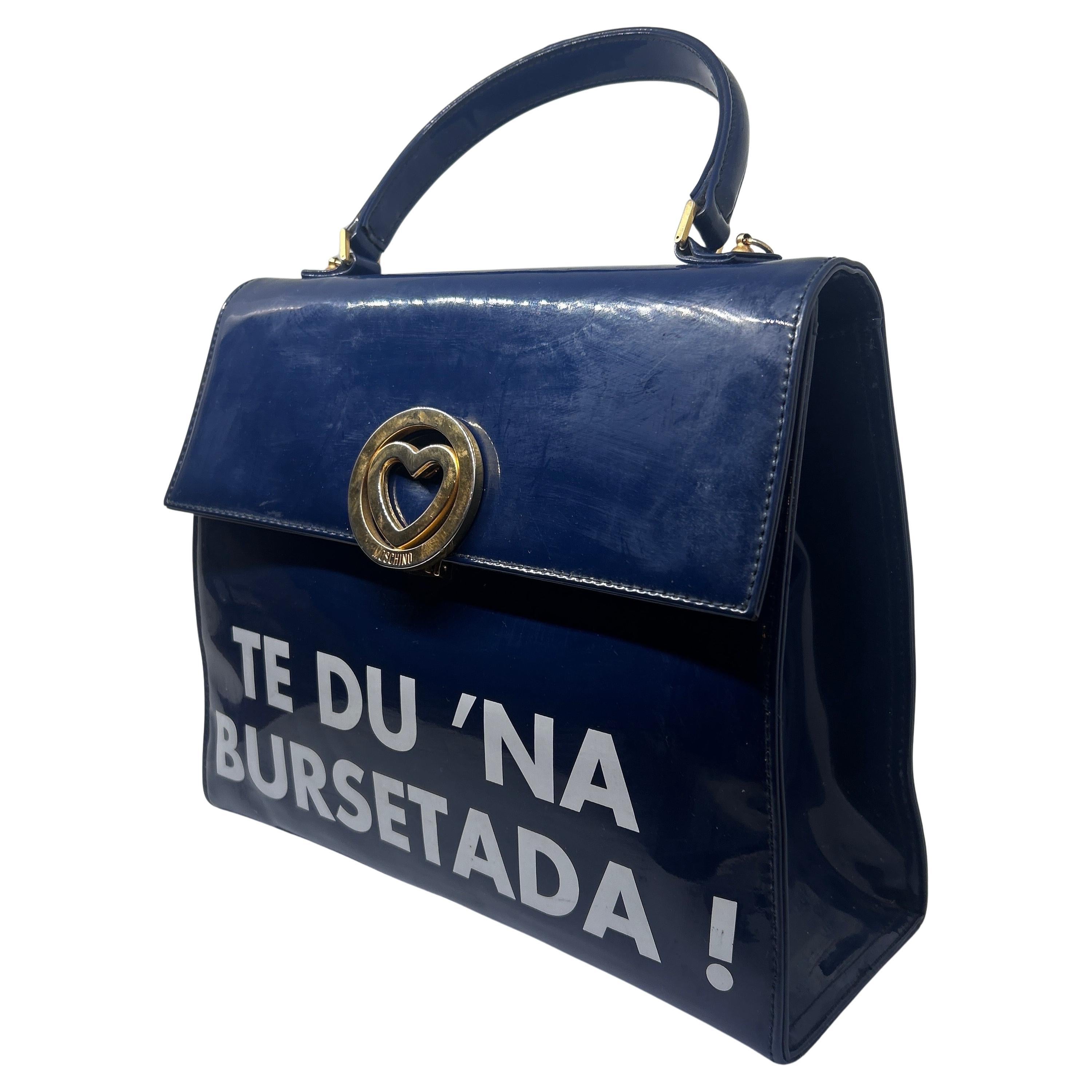 This piece exemplifies Moschino's characteristic sense of humor and playful approach to fashion.
The bag is a structured, dark blue plastic handbag with a top handle. It has a bold statement printed in white lettering on the front that reads 