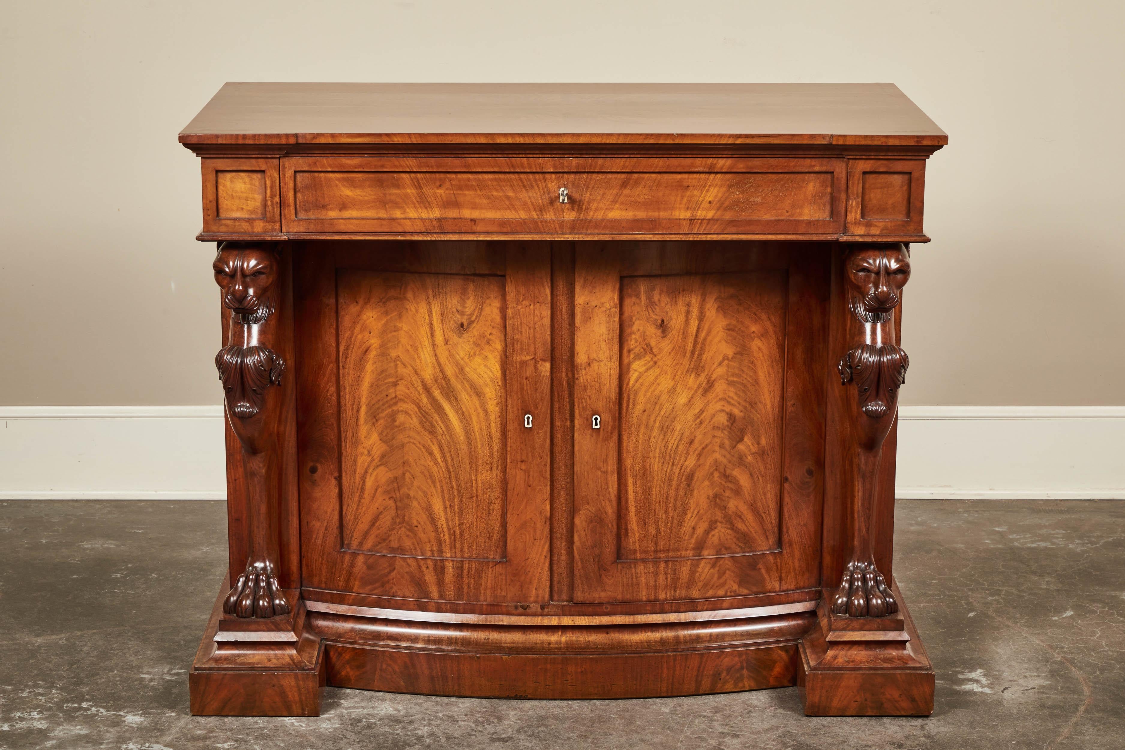 A William IV style mahogany secretary cabinet. Outset surface with a molded edge over a single drawer which opens to reveal a writing surface and three small drawers, set above a bowfront two door cabinet flanked by carved griffin monopedia