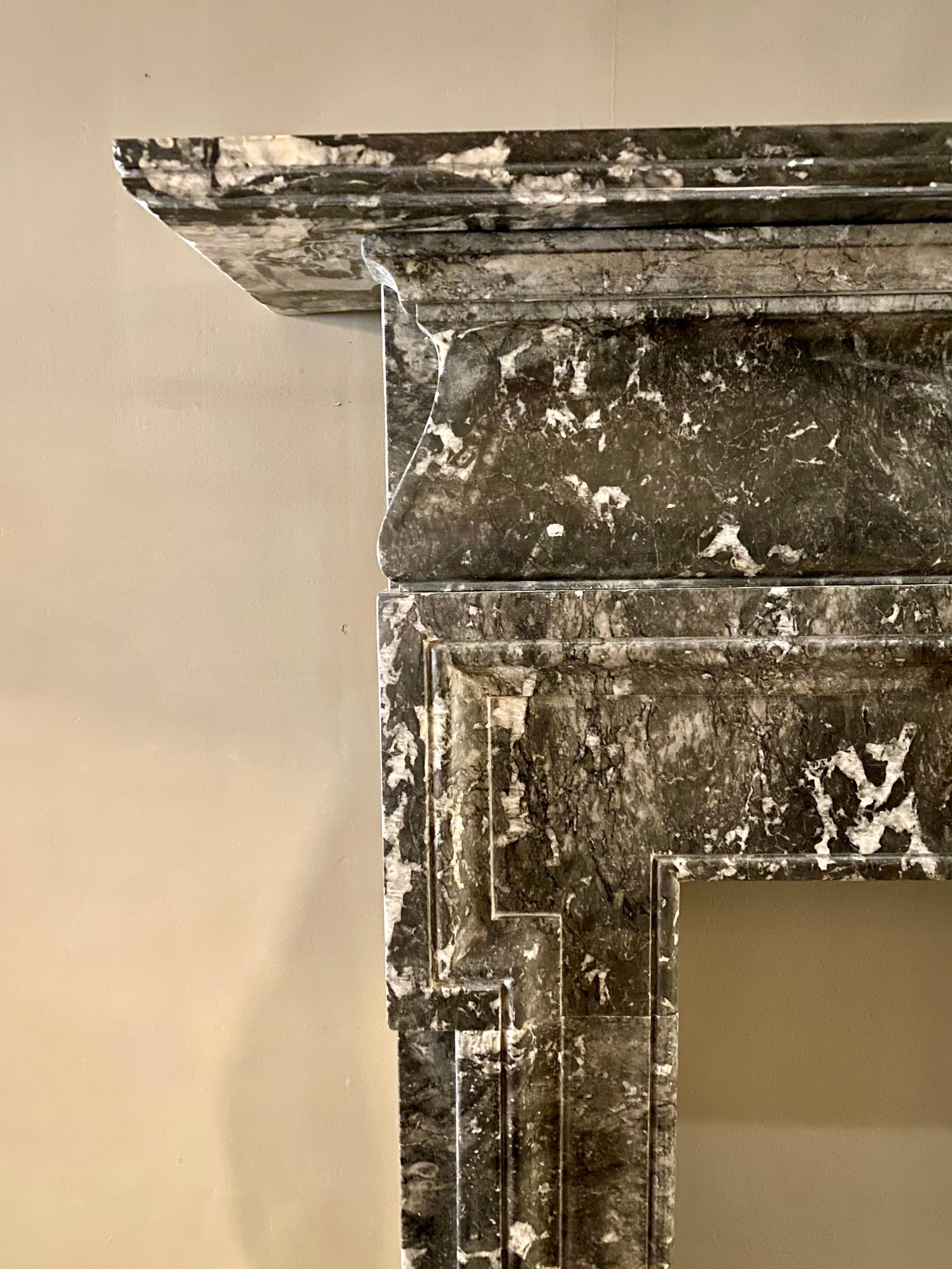 A continental fireplace in a variegated Grey Fossil marble. Architectural in design with, simple eared surround. The frieze with a central fielded plaque, flanked either side by scrolled ended undulated panels, with stepped moulding above. All