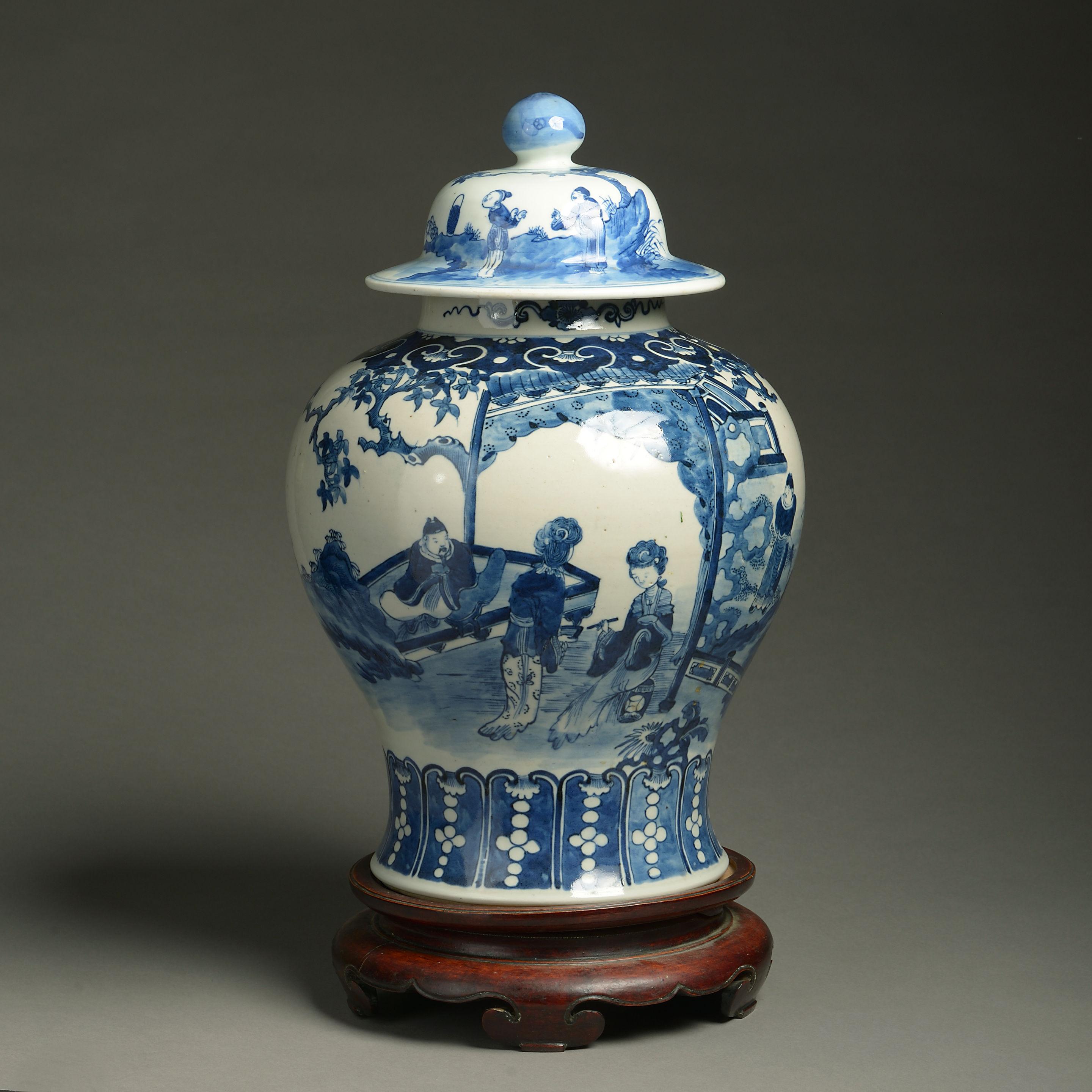 A late 19th century blue and white glazed baluster form vase and cover, decorated throughout with courtly figurative scenes. 

Late Qing dynasty. 

Together with a carved turned wooden stand. 

Dimensions listed are excluding the base.