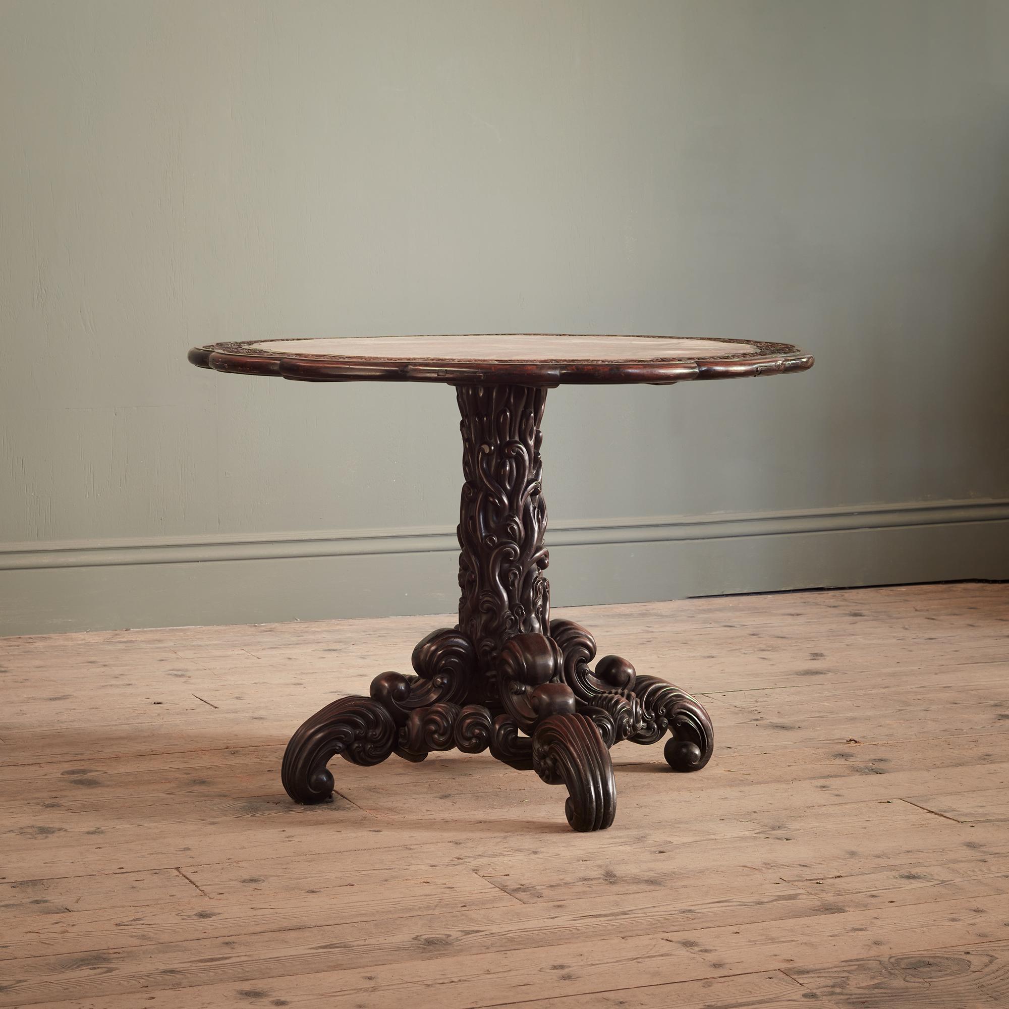 Tiene forma lobulada circular con encimera de mármol rosa incrustado, sobre una base de pedestal tallada con textura y una elaborada columna central.