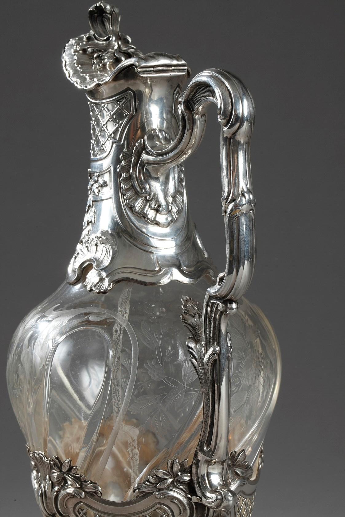 A crystal ewer blown and carved with facets in silver mounts. The crystal is engraved with flower baskets or baskets of flowers. The neck of the ewer is finely chiseled with floral motifs and grape vines. The beak is closed by a lid whose grip is