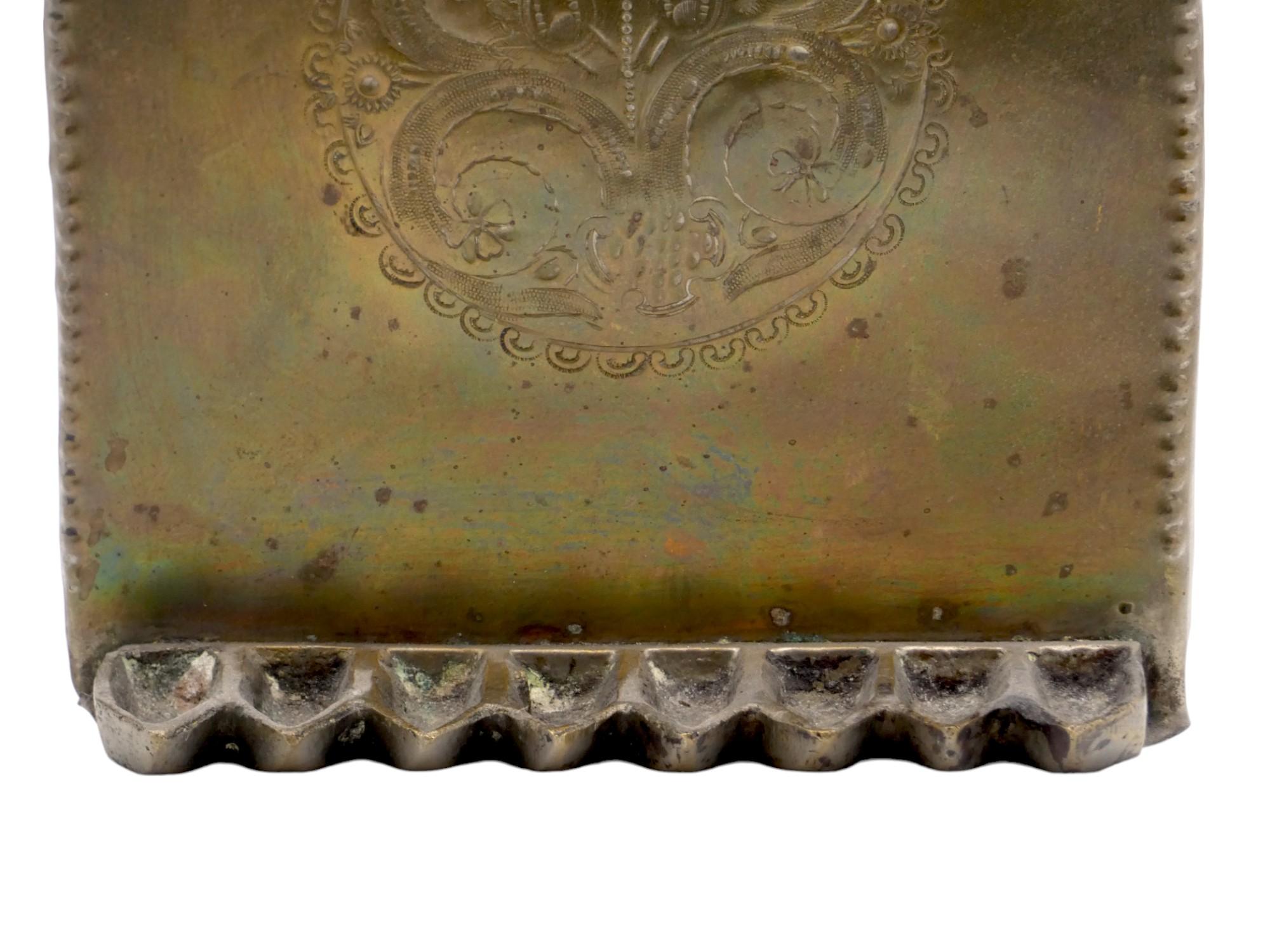 Un'insolita lampada di Hanukkah in ottone del XIX secolo.

Questa lampada di Hanukkah ha una forma insolita, con una cornice di perline e una piastra posteriore alta con un cartiglio centrale delimitato da volute a C e una saporita decorazione