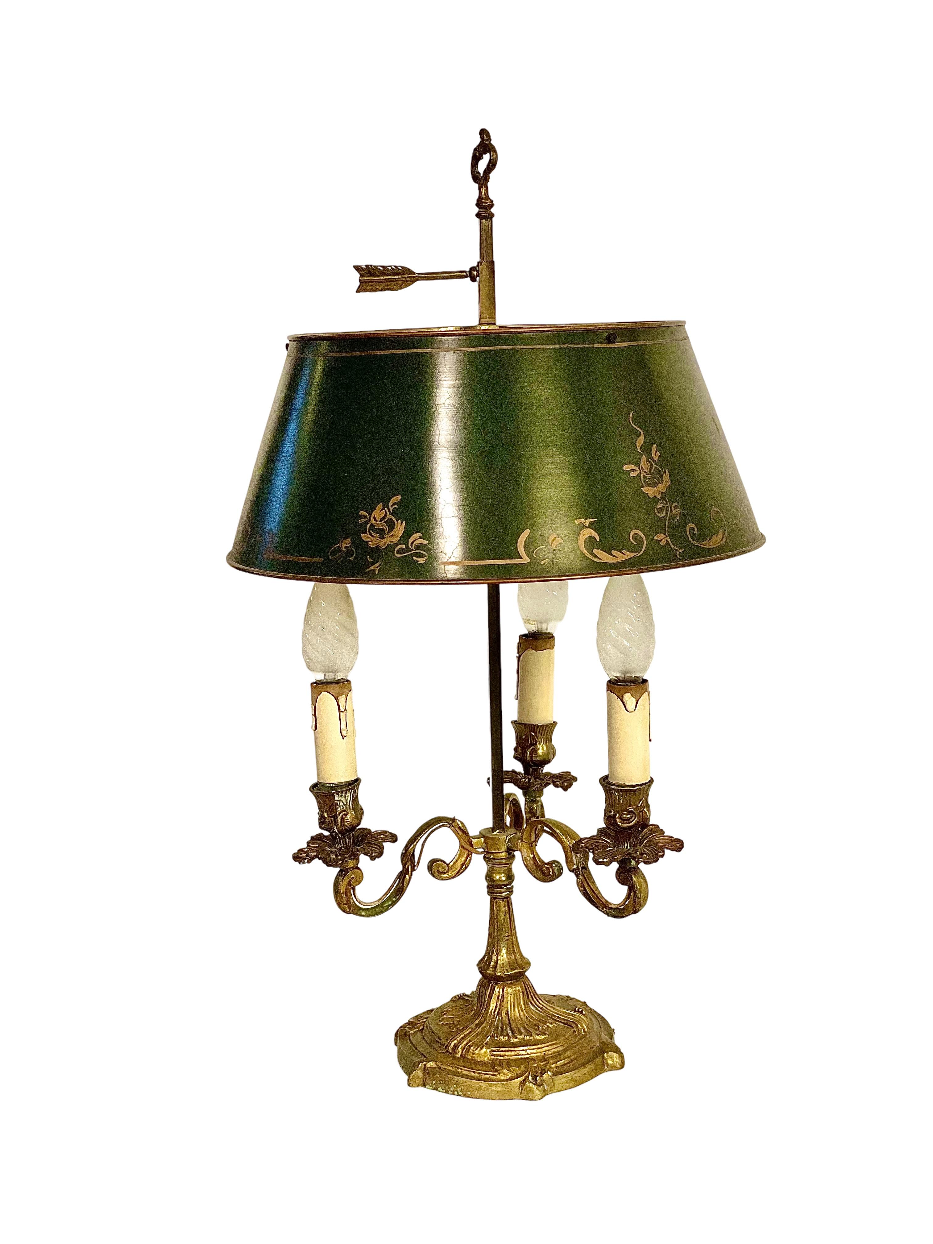 A late 19th century Louis XV-style Bouillotte table lamp, with original, green, gilt- decorated tole lampshade, above a base of three upturned decorative brass candle arms supported above a heavy base. Featuring a tall carrying handle with a festoon