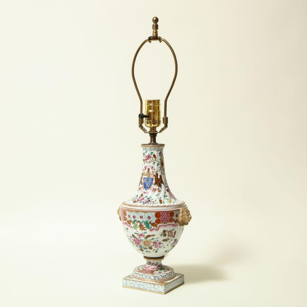 Conceived in the Chinese export style; the base of urn form on a sole base and with lions' masks applied to the side; profusely decorated with polychrome sprays and bearing an apocryphal armorial; fitted with a brass light socket and removable harp.