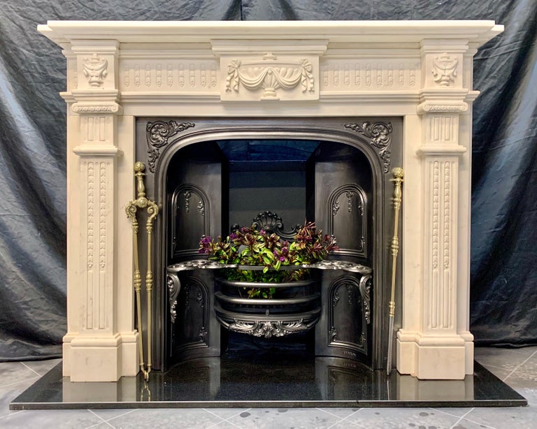 A 19th century Georgian style statuary marble fireplace surround of good proportions and color, a profiled top shelf rests above a central tablet displaying a large urn, flanked by frieze panels of vertical flutes and bell flowers, further urns to
