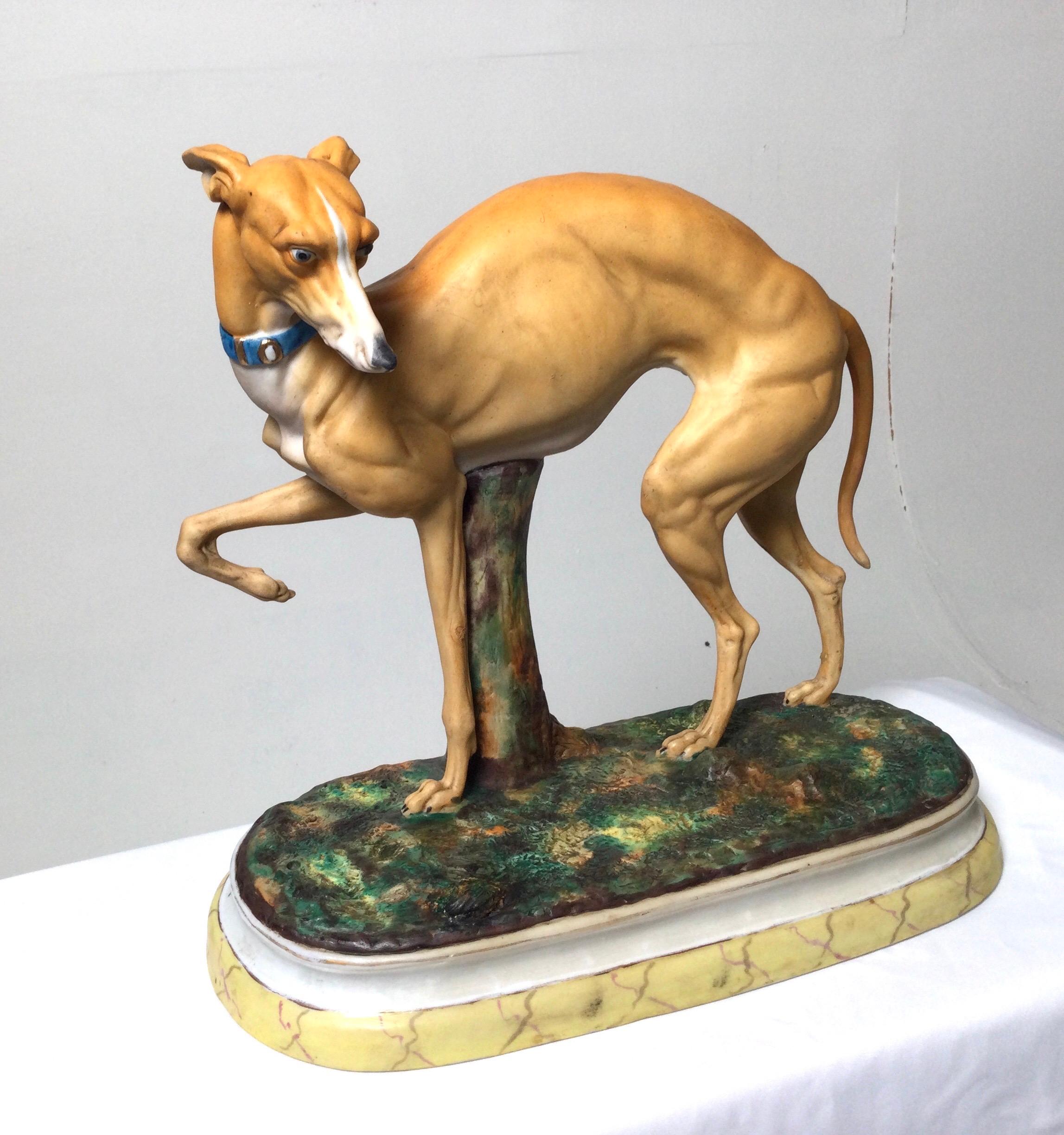 Graceful porcelain figure of a whippet dog. The 19th century sculpture with a hand panted bisque surface depicting dog with blue collar on a patch of variegated green and brown grass. The oval base with a yellow marble paint decoration. Beautifully
