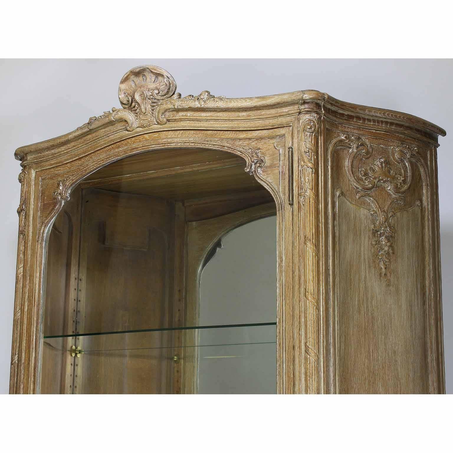 A fine 19th century Louis XV style country French - Provincial carved white-washed walnut bombé credenza vitrine. The slender and tall carved single-door vitrine with a bowed front glass door and three adjustable glass shelves, crowned with a carved