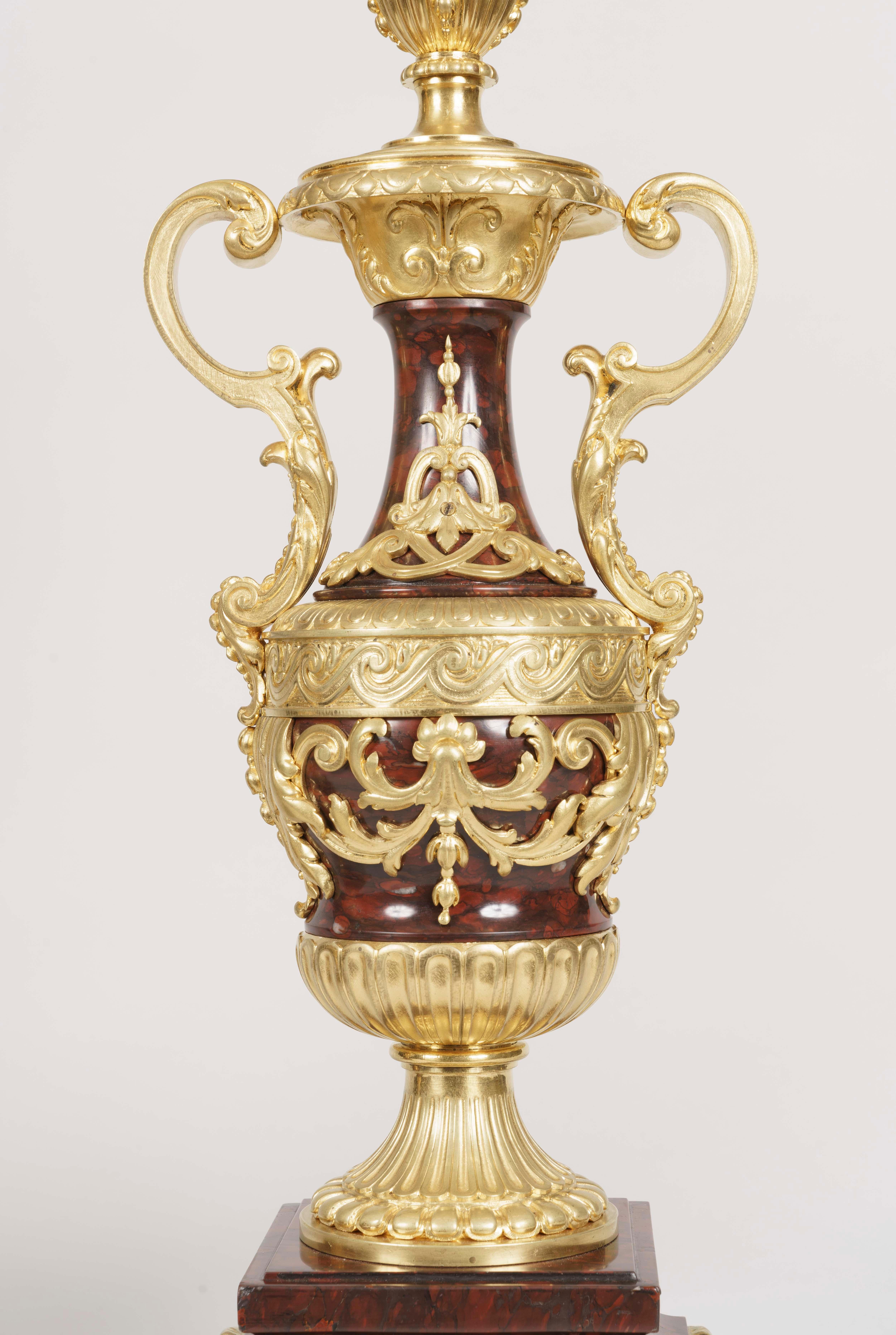 A pair of Louis XVI style marble and gilt bronze candelabra.

Rising from a square base supported by gilt bronze scrolling lions' paw feet, the red 'partridge eye' griotte marble sculpted in the form of a Greek Loutrophoros vase, the scrolling