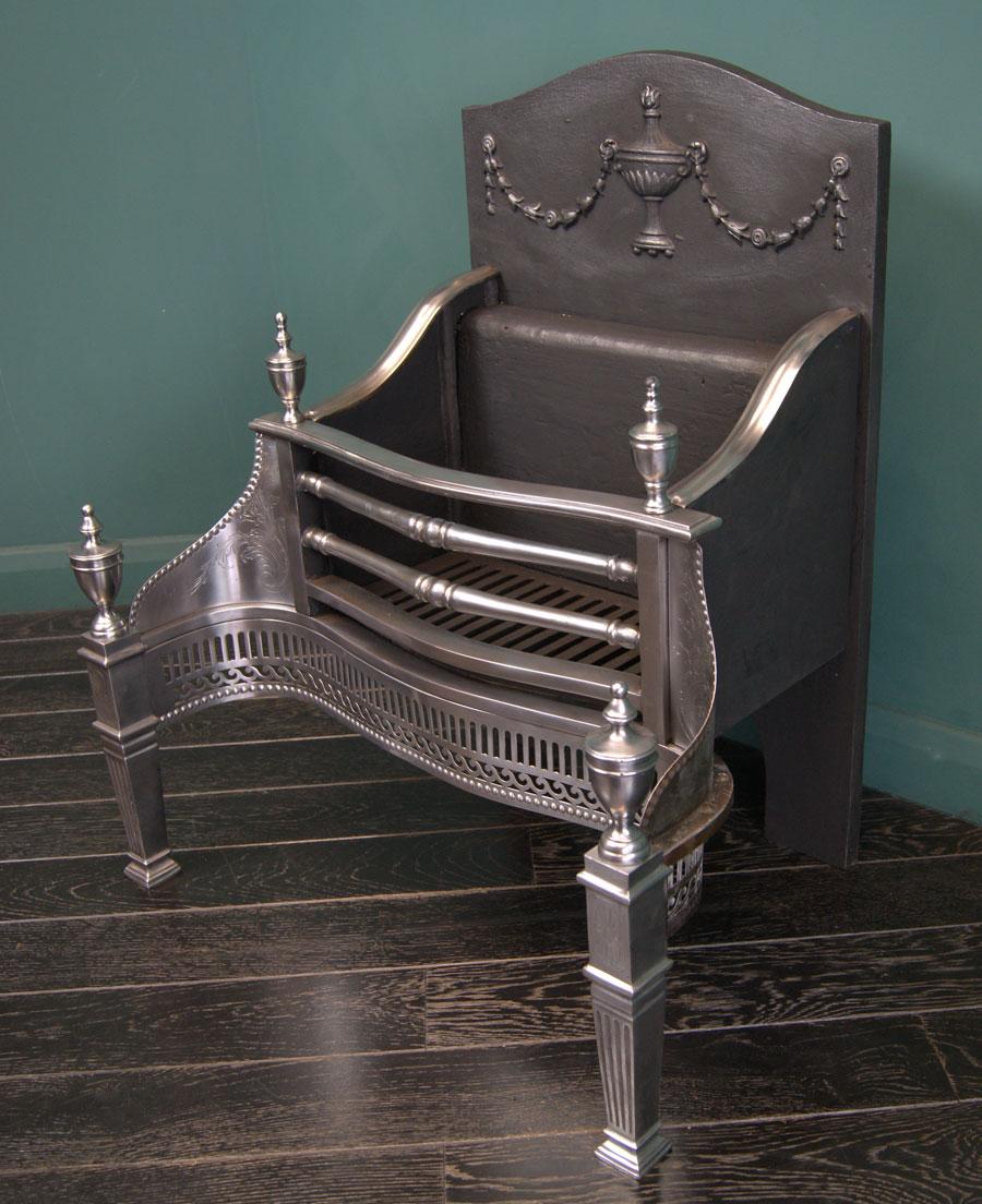 Grille de foyer robuste en acier poli en parfait état. La façade en acier poli avec des barres de feu ornées, flanquée d'ailes gravées de feuillages sur une frette cannelée et à volutes. Des épis de faîtage en forme d'urne sont placés en haut devant