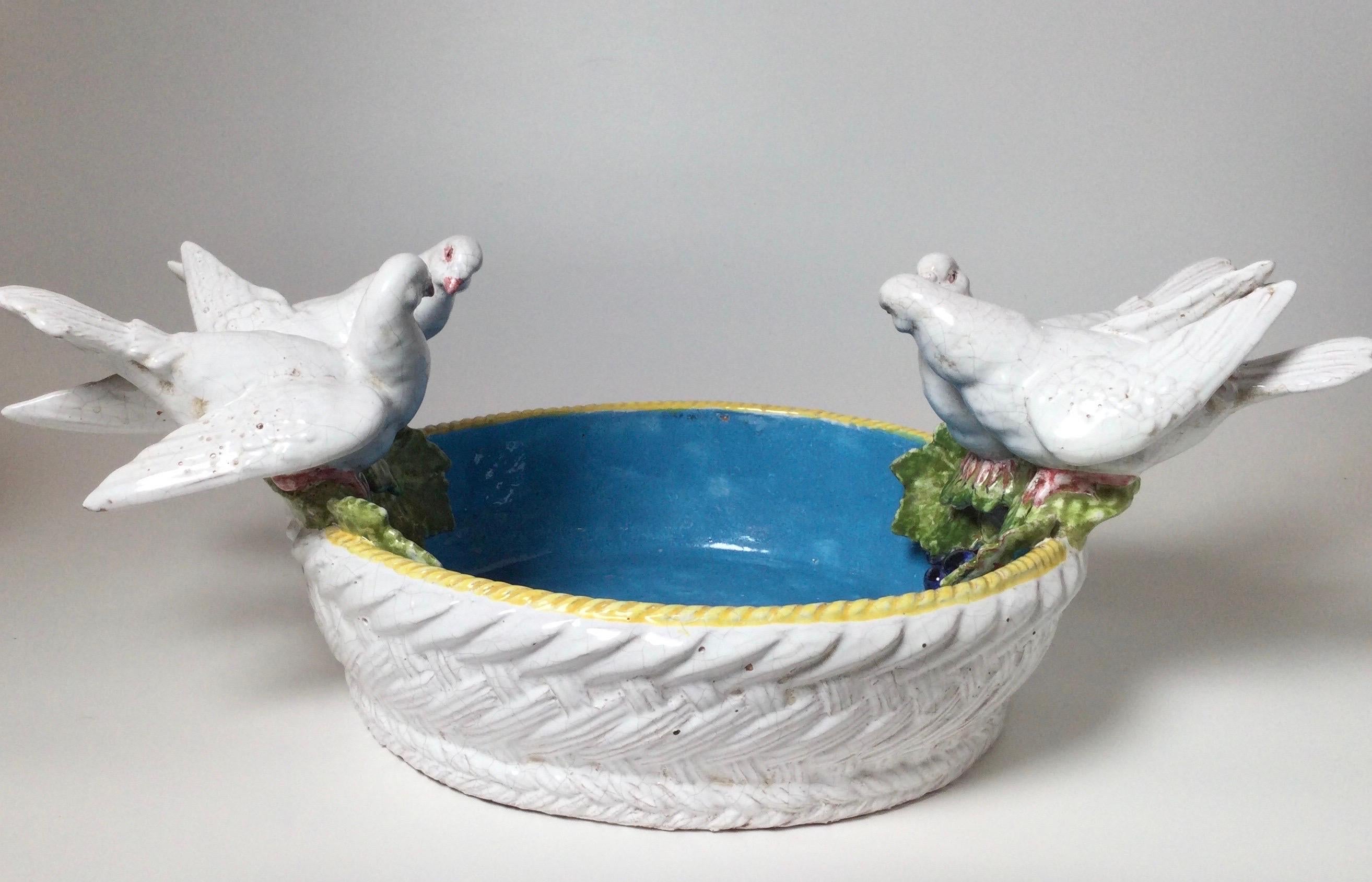 A whimsical large open bowl in the form of a bird bath. The white doves perched on the rim of a bowl with an azure blue interior. The bowl in a basket form with grapes and leaves. Vibrant color on white faience.