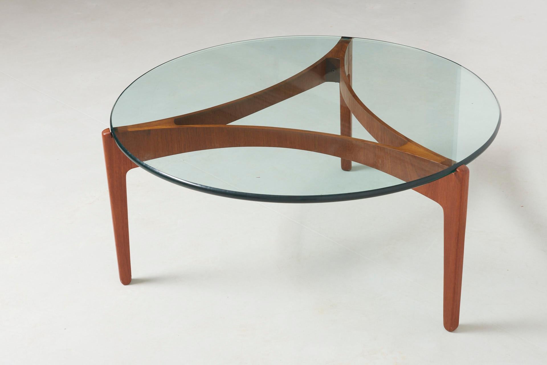 A round low table with glass tabletop resting on a three-legged base. Design by Sven Ellegaer for Christian Linneberg. The legs are made of teak. The glass top has a nice rounded edge.