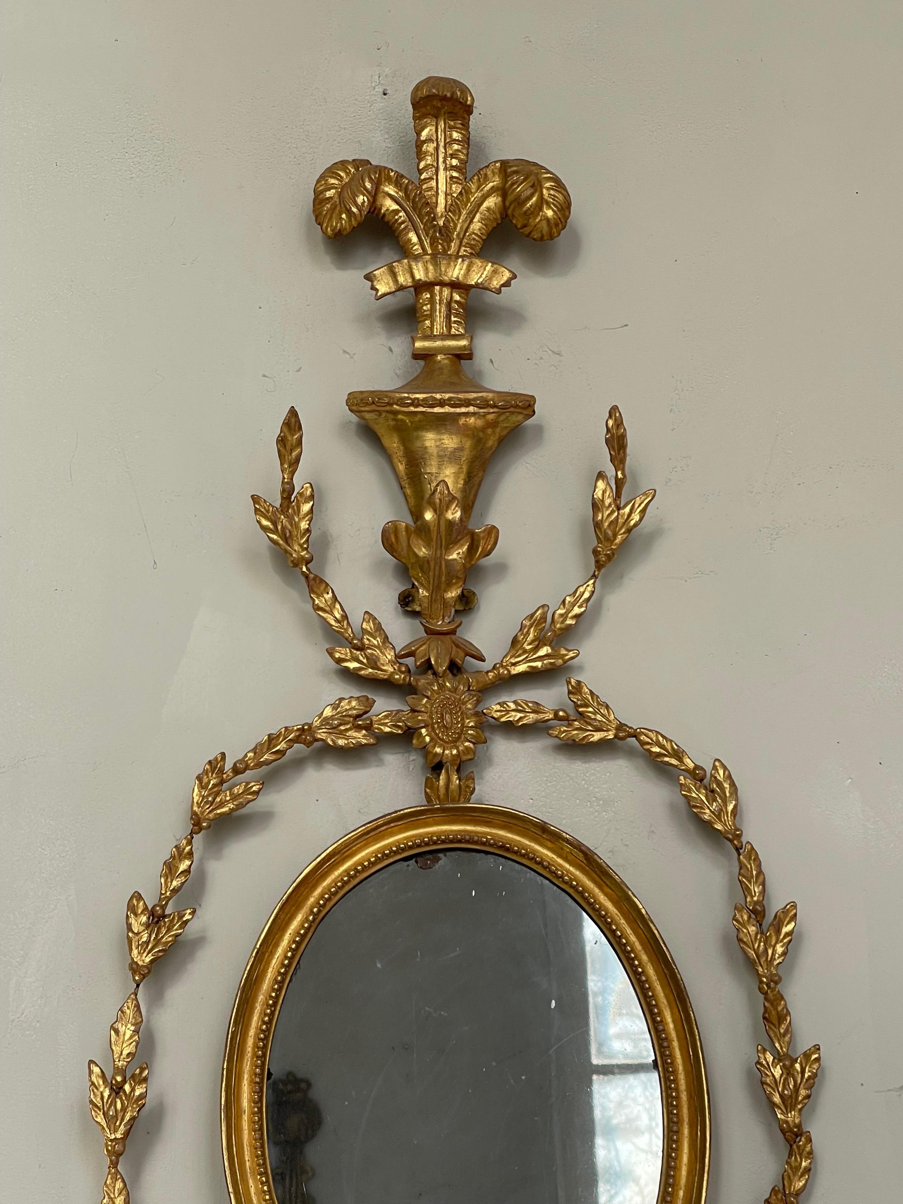 This extremely rare pair of giltwood girandole mirrored wall sconces is English and was produced in the early 19th century in the Adam style.
A bracket supports a foot tall oval framed looking glass surrounded by intertwined Laurel swags. Then from