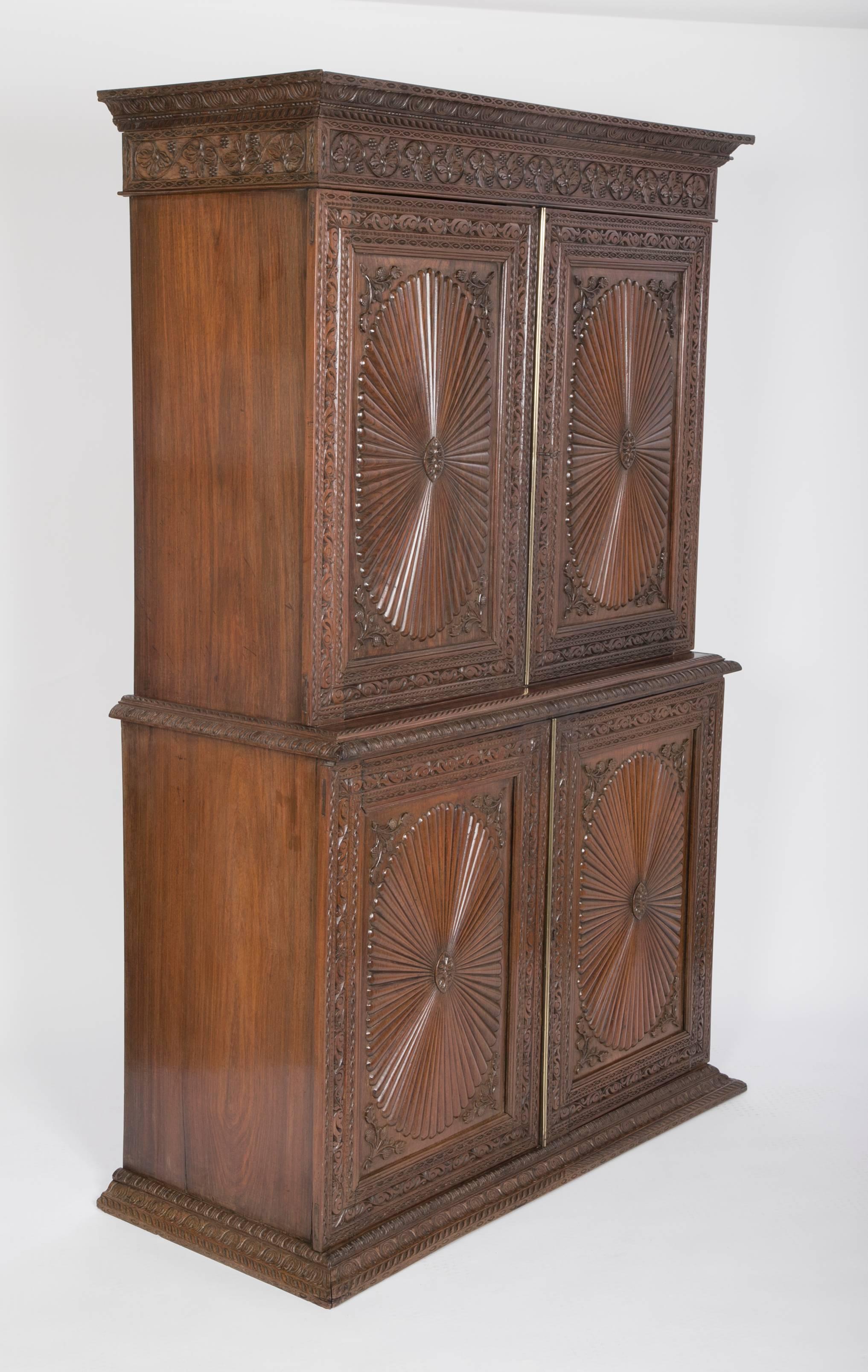 A mid-19th century, Anglo-Indian linen press with four carved stylized sunburst paneled doors and floral carving through out. The upper and lower case each with two shelves.