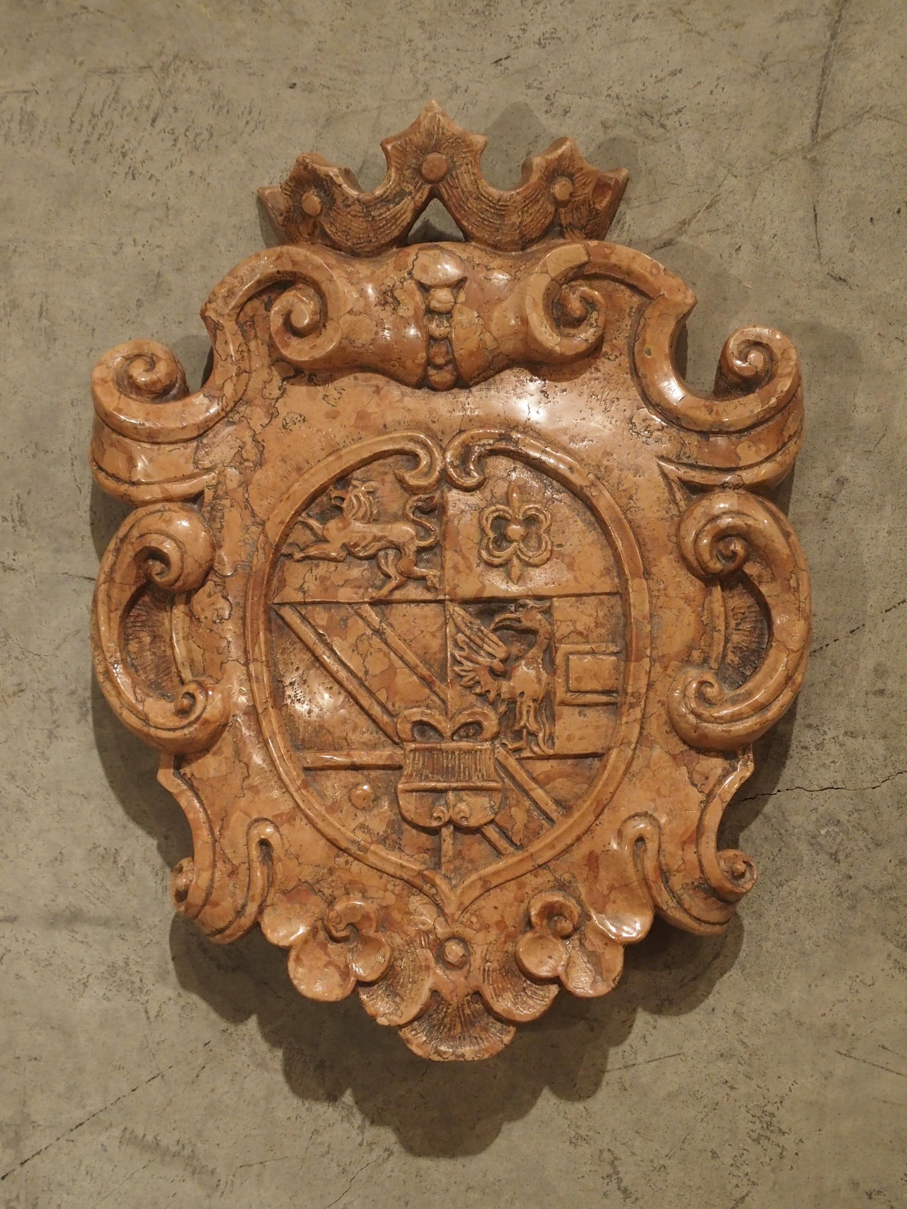 This shield has been carved out of one solid piece of Rosso Verona marble. A stylized crown is at the top of the shield, which is surrounded by a series of different scrolls with floral motifs. In the central coat of arms section, we see images of a