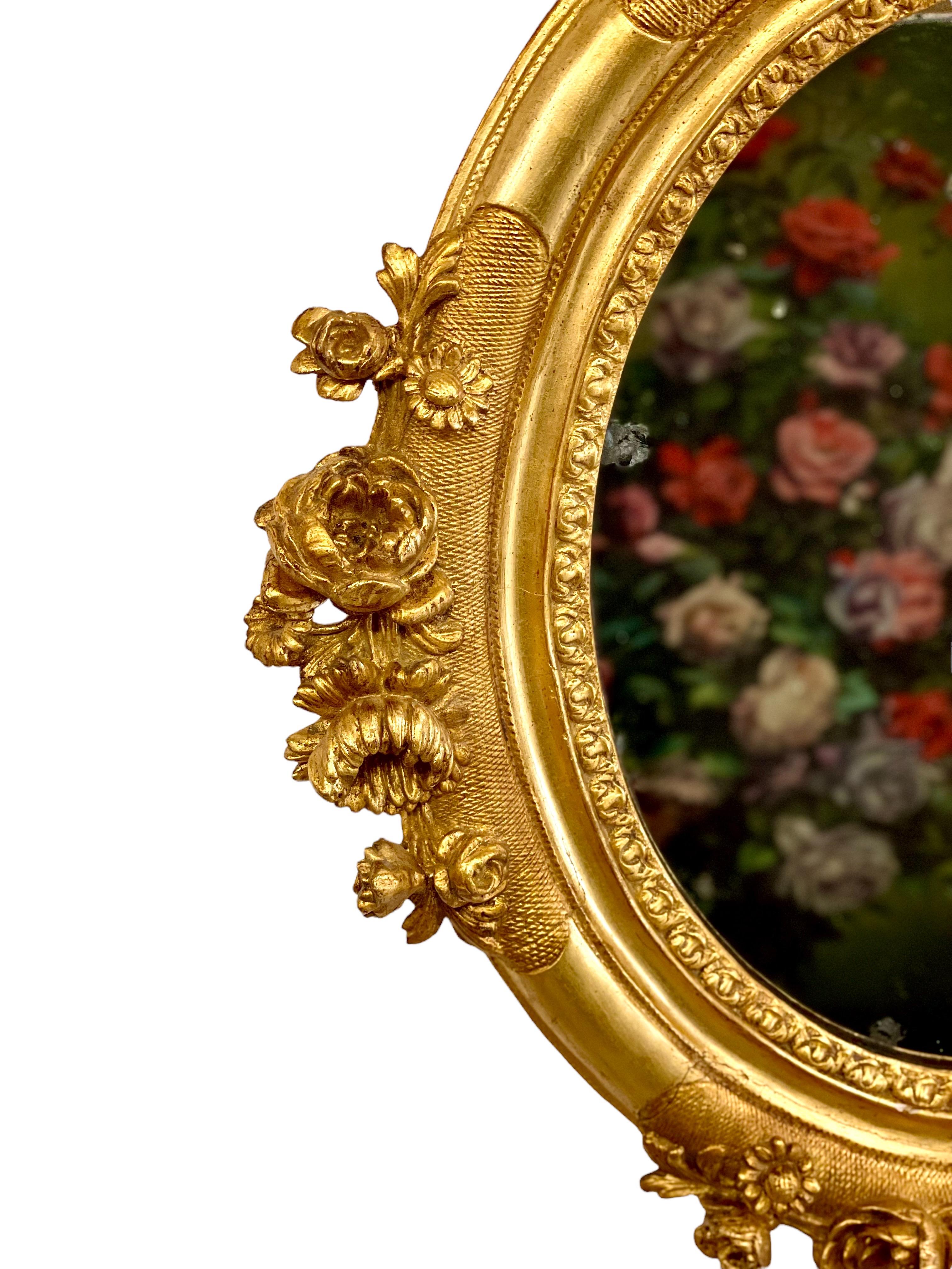 A beautifully carved gilt wood and stucco framed Louis XVI style oval mirror, embellished with floral motifs of paeonies, daisies and chrysanthemums in high relief on its four sides. This elegant mirror would be a striking adornment on a bedroom