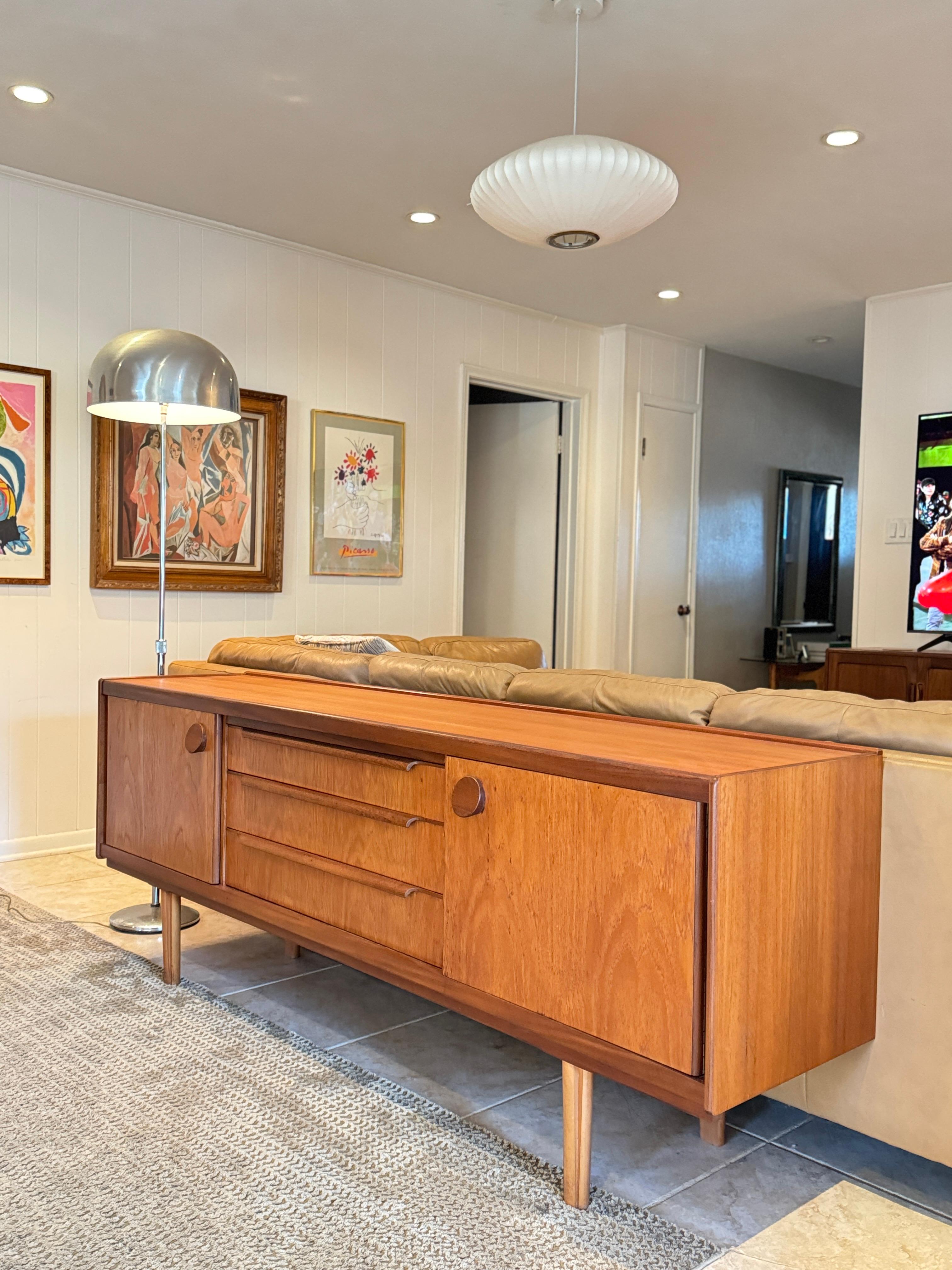 Una credenza in teak di metà secolo, circa anni '60, dal design accattivante. Con pomelli tondi in teak massiccio per i cassetti e maniglie a pinna per le ante, due armadi a ripiani di buone dimensioni e tre ampi cassetti centrali. Il teak ha una