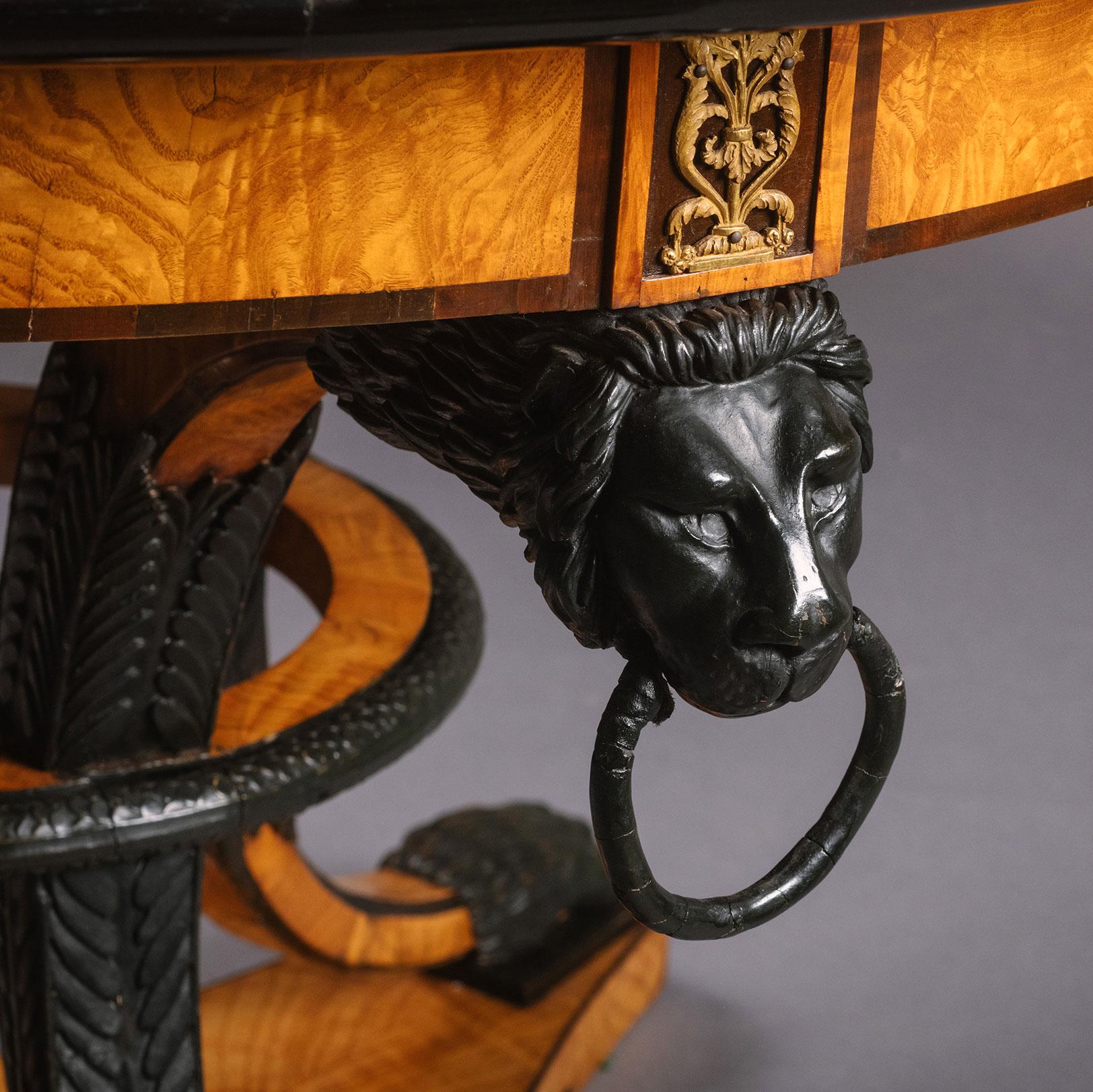 A Biedermeier Satin birch and ebonised centre table.

This handsome centre table has a circular top of beautifully figured birch. It rests on sabre-shaped legs headed by lion-masks holding rings and joined by a loop stretcher modelled as a snake