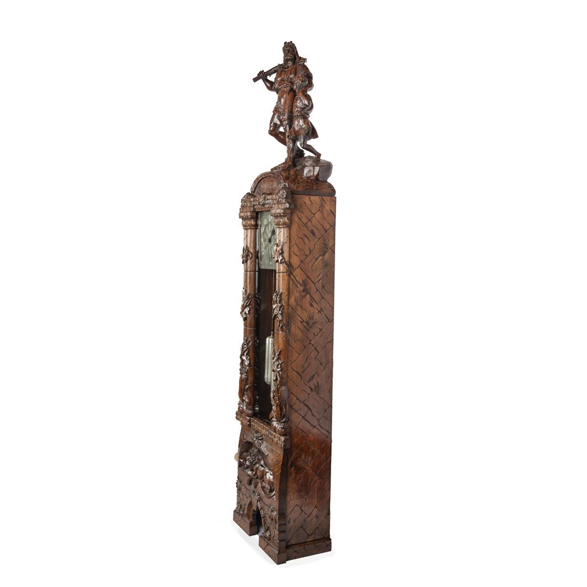 A ‘Black Forest’ linden wood long case clock by Spring of Interlaken, the square silvered dial enclosed behind a glazed door within an ornate case, surmounted by a carving of William Tell and his son after the “Tell Monument” by Richard Kissling