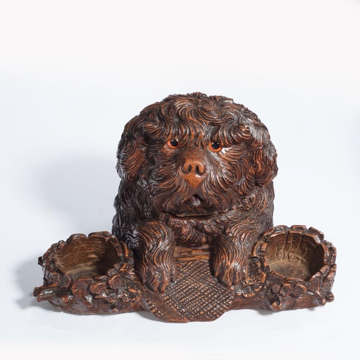 A ‘Black Forest’ walnut tobacco box in the form of a long-haired dog resting its forepaws on a log carved with a well at each end, the head, naturalistically carved with curly hair and inlaid glass eyes, is hinged and opens to reveal a hollowed out