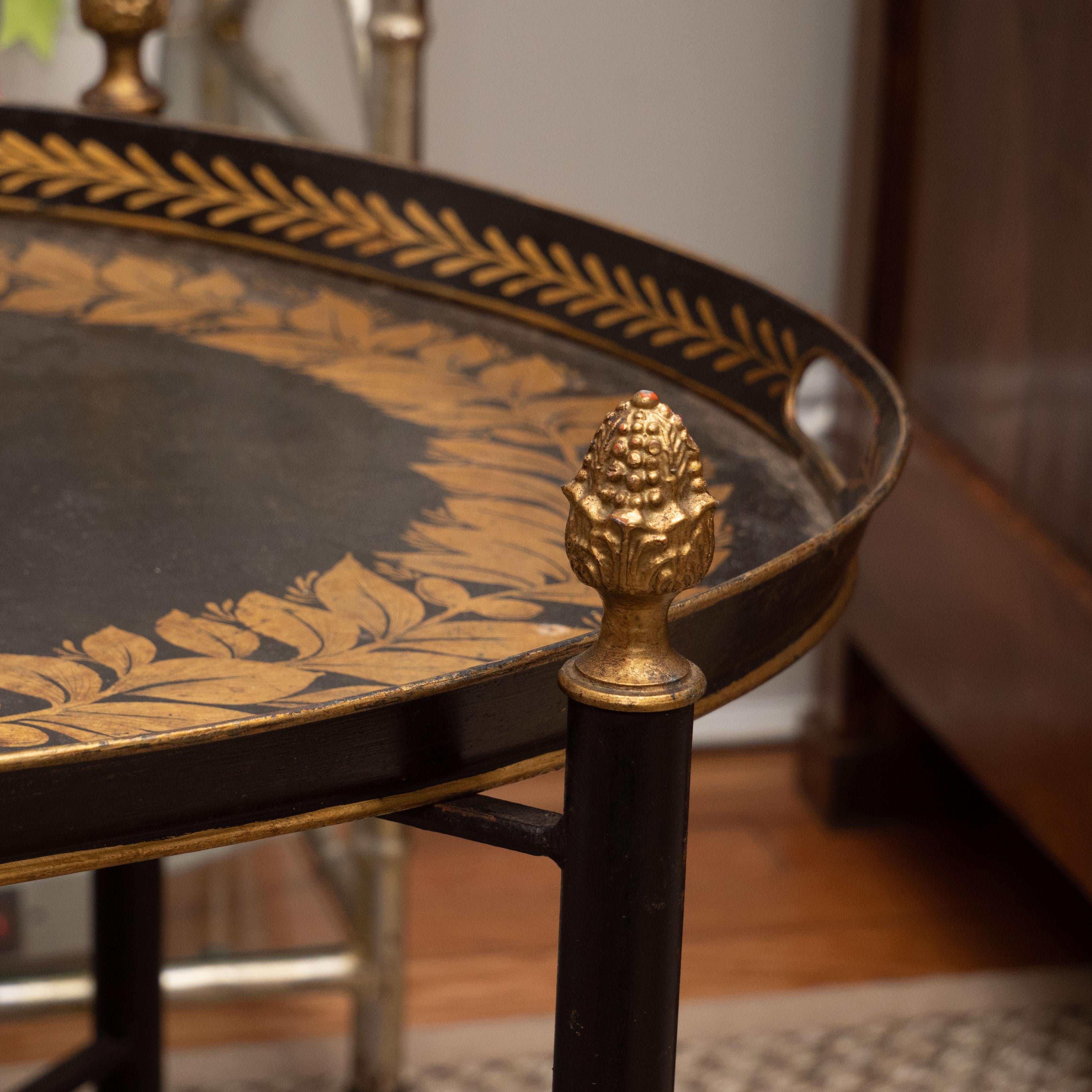 A lovely oval tole tray on a custom stand in black with a hand painted design in gold. The X-form stand features charming pine cone finials. This table is perfect as a side table or between two chairs. Without taking up much space, it adds style and