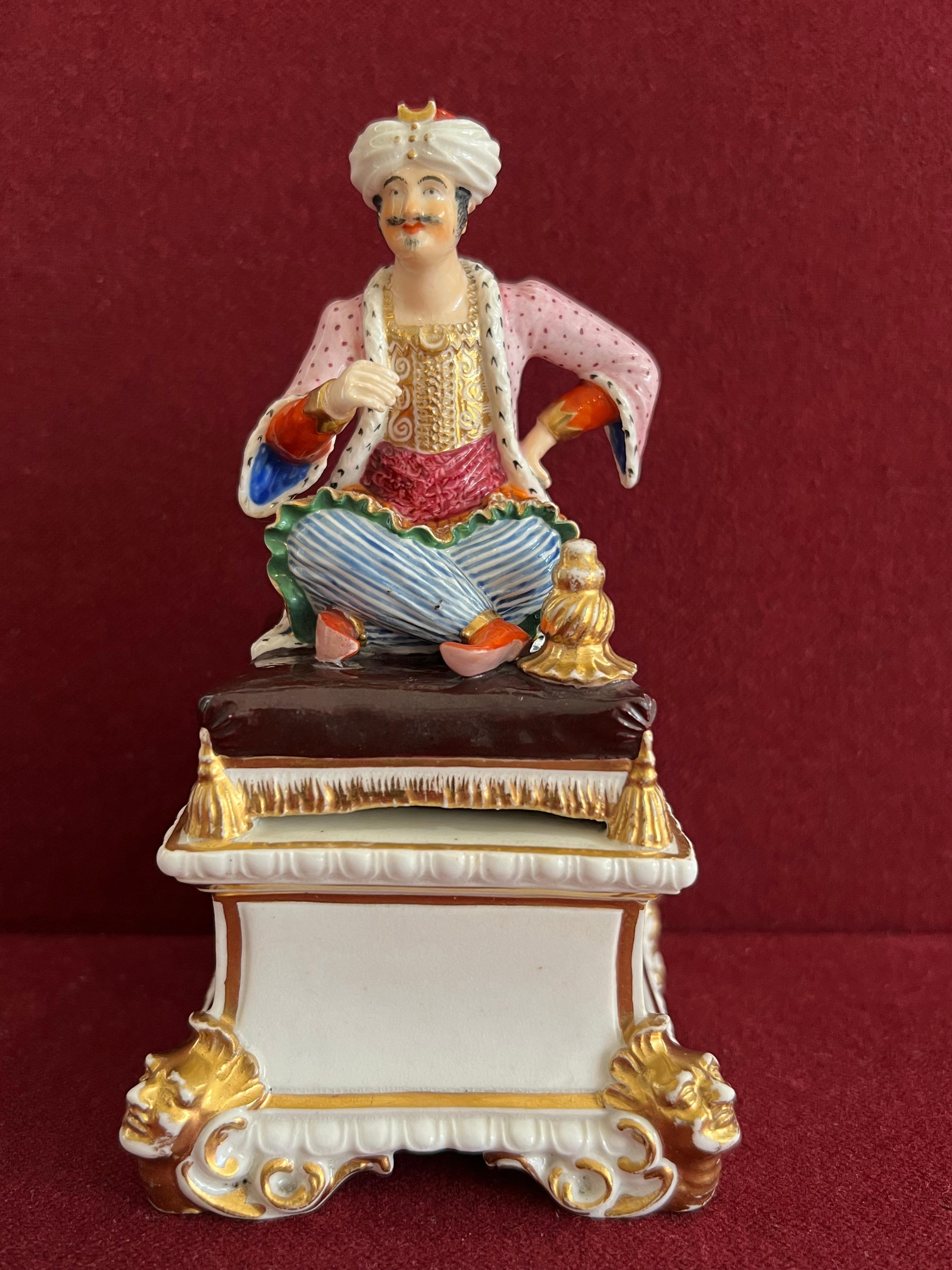 A very fine Bloor Derby porcelain figure of a seated Turk, sitting cross-legged in traditional costume, a hookah at his side, on a cushion base and square plinth with mask corners. Brightly decorated in enamels and gilding. Crown and Bloor Derby