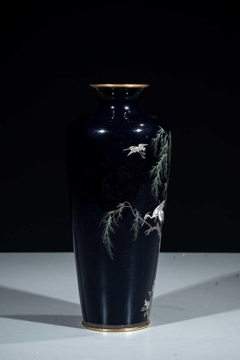 A cloisonné vase decorated on a dark blue ground with a pair of herons perched on the branch of a weeping willow and another heron in flight.

The decoration is executed with silver wires of varying thickness and multicolored enamels.

Period: