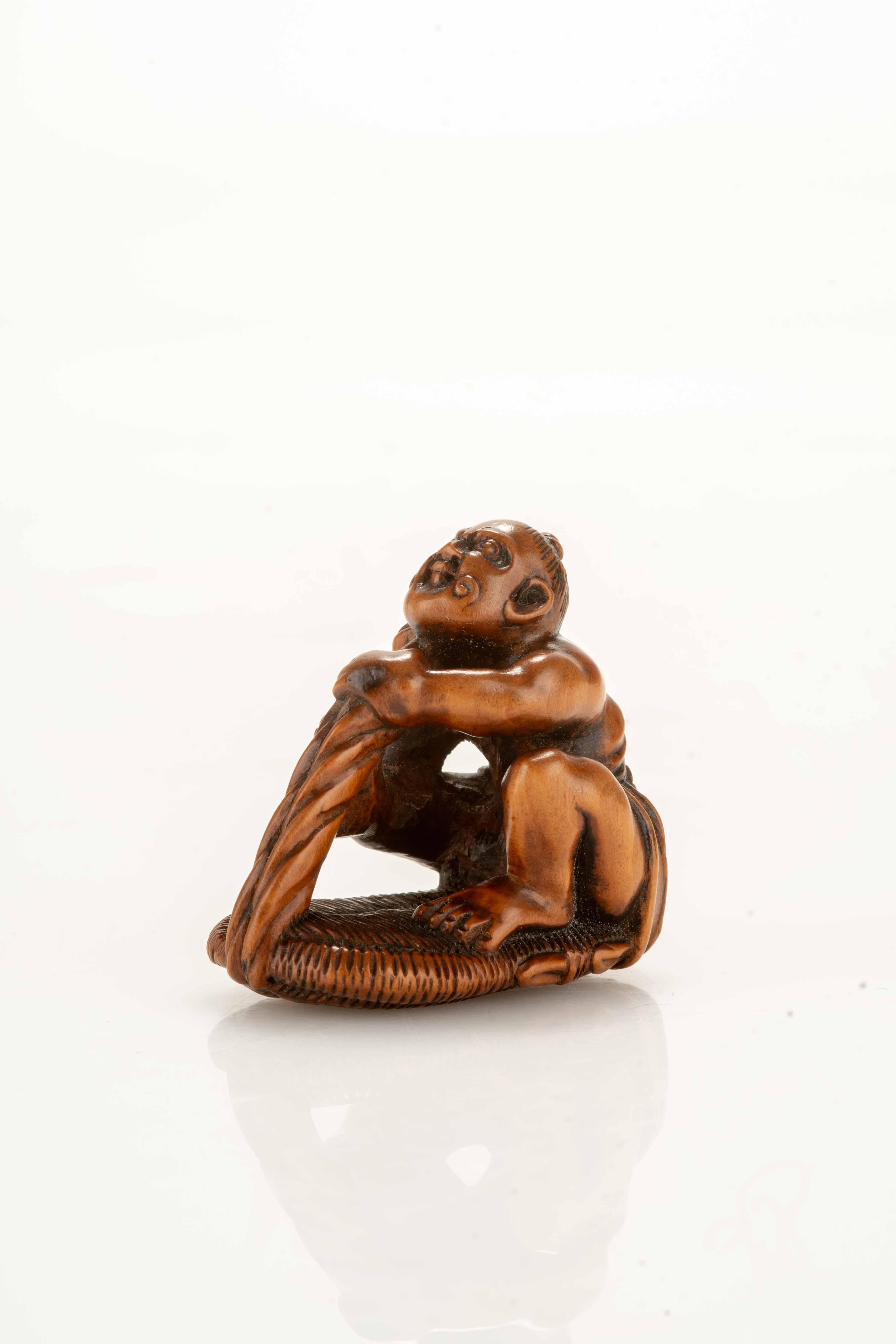 Boxwood netsuke depicting a Nio engaged in braiding the strings of a giant sandal. The Niō's expression is intense with sculpted muscles in tension and a determined face. Ringed Himotoshi and Hokei signature engraved under the base with a horn