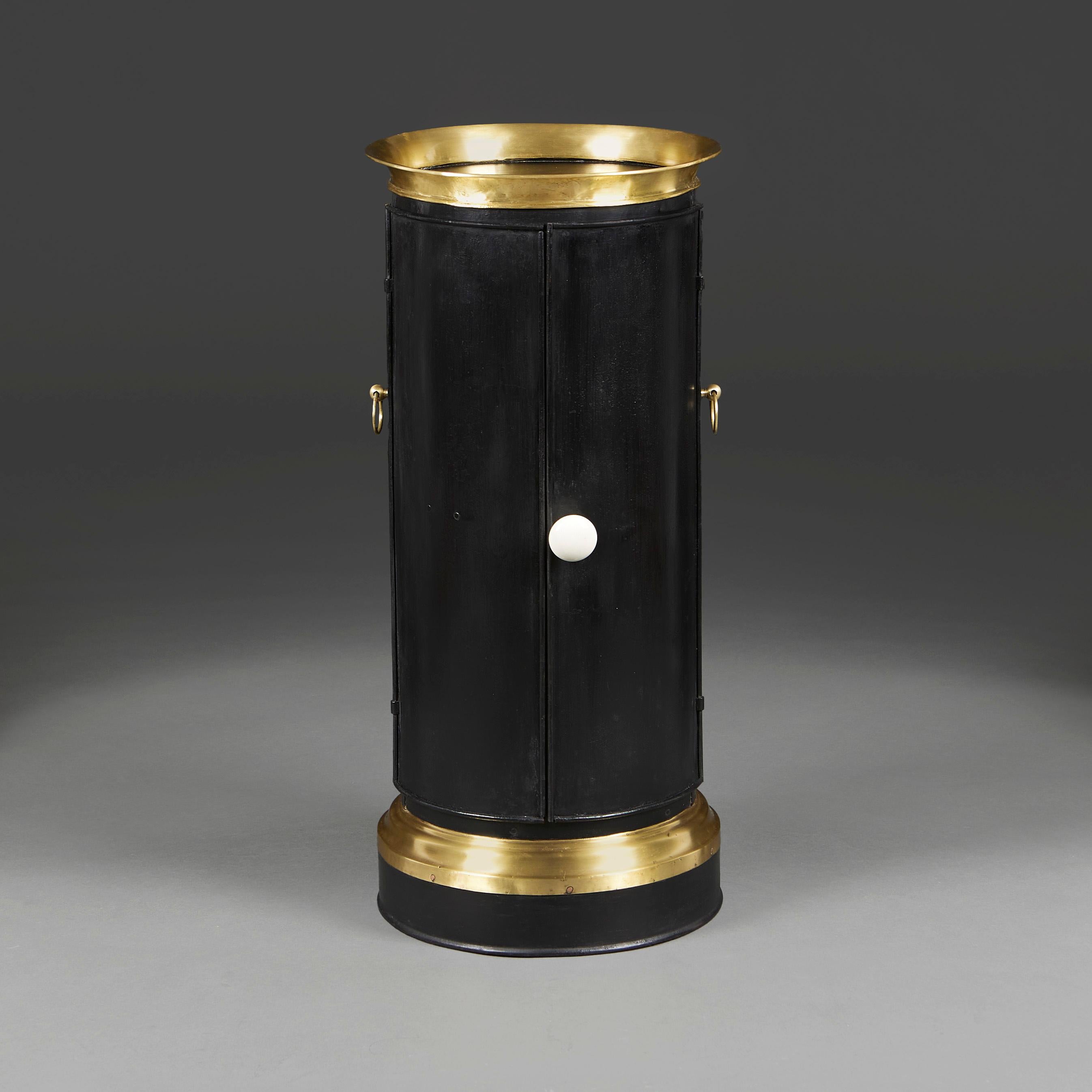 An unusual late nineteenth century black metal and brass cylindrical cabinet, opening with two doors to the front to reveal three interior shelves, with brass banding to the base and a solid brass out swept gallery to the top, with brass loop