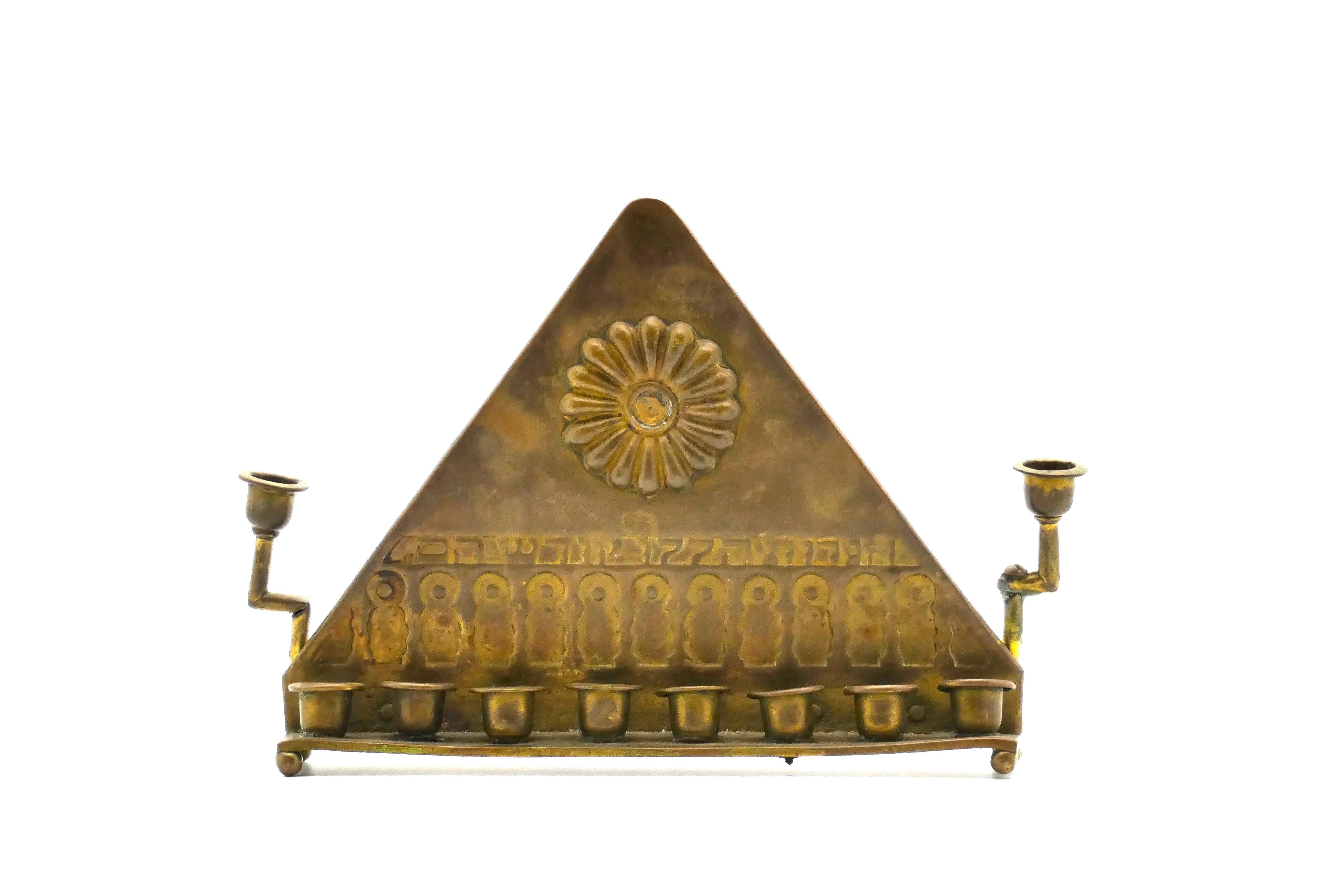 This Bezalel Hanukkah lamp is elegantly decorated with a prominent rosette on its triangular backplate.

On the lower half of the backplate are numerous notched archways placed under the similarly indented inscription in Hebrew which translates to