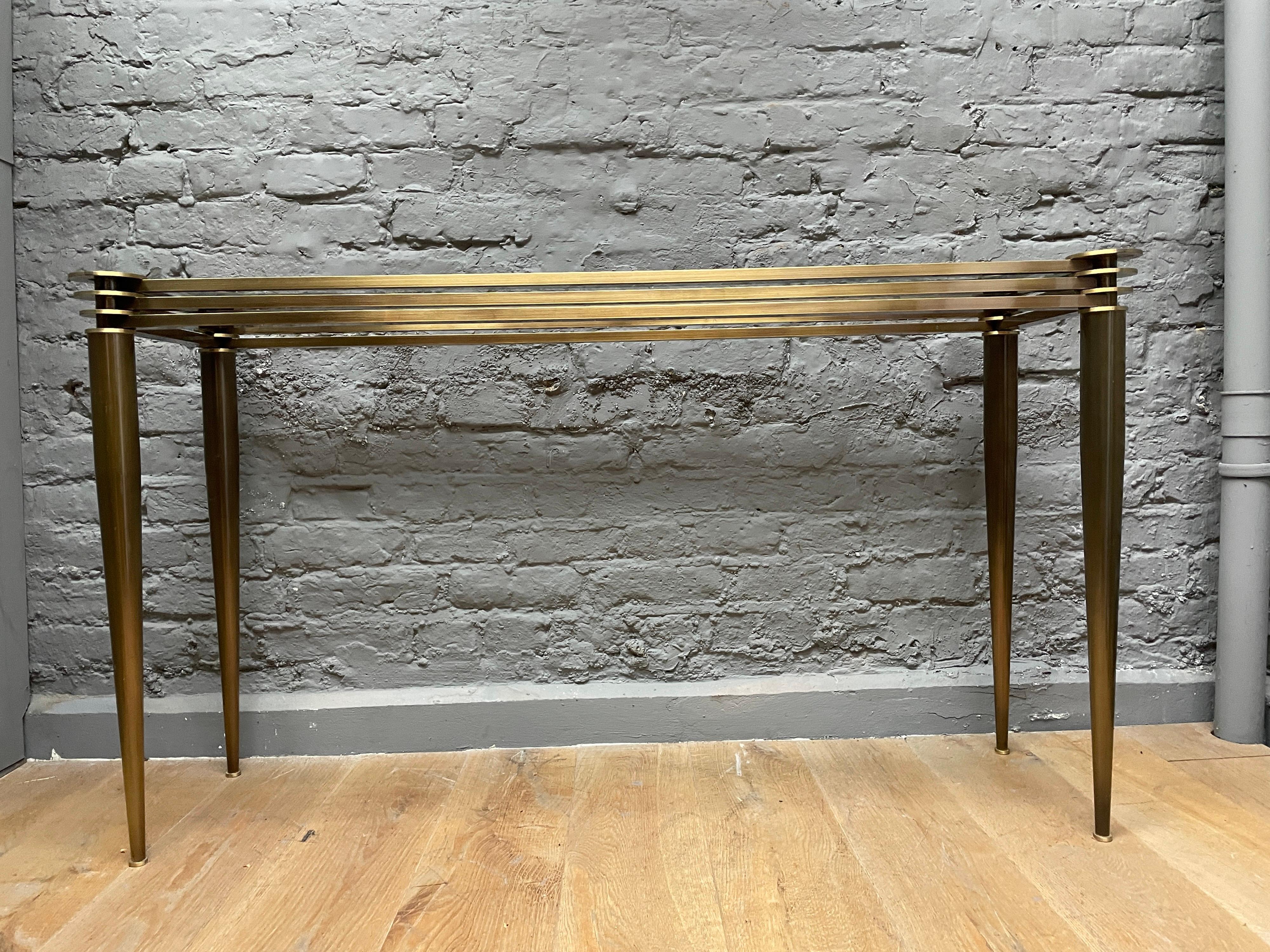A midcentury style console table in brass and patinated brass/bronze finish. The conical tapering brass patinated supports on circular brass feet terminating in 4 ascending brass rings with stepped banded brass supports holding the glass top.