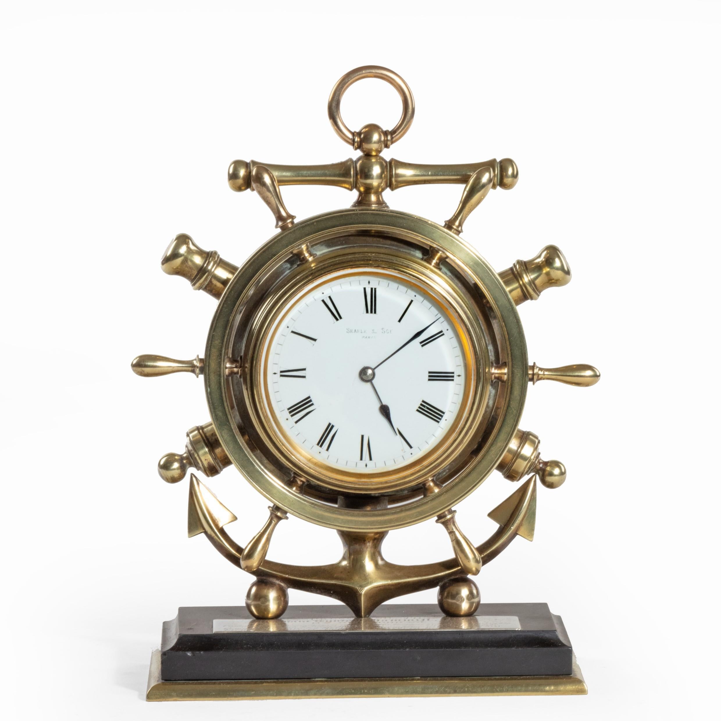 A brass ship’s novelty clock presented to Captain Tynte F Hammill RN., in the form of a ship’s wheel set against an anchor and crossed cannon, all raised on cannon ball feet, on a wood and brass base, inset with a silver plaque reading; Presented to