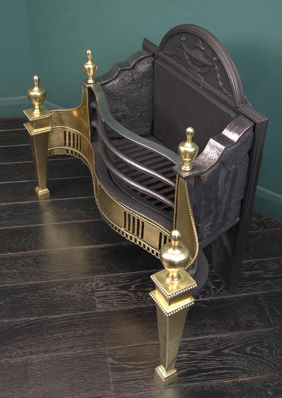 A large English Adam-style polished brass fire grate (by Thomas Elsley). The railed serpentine basket above a fret with interrupted flute piercing is set between square tapered legs with large brass urns. Engraving to centre-apron. Wrought-iron fire